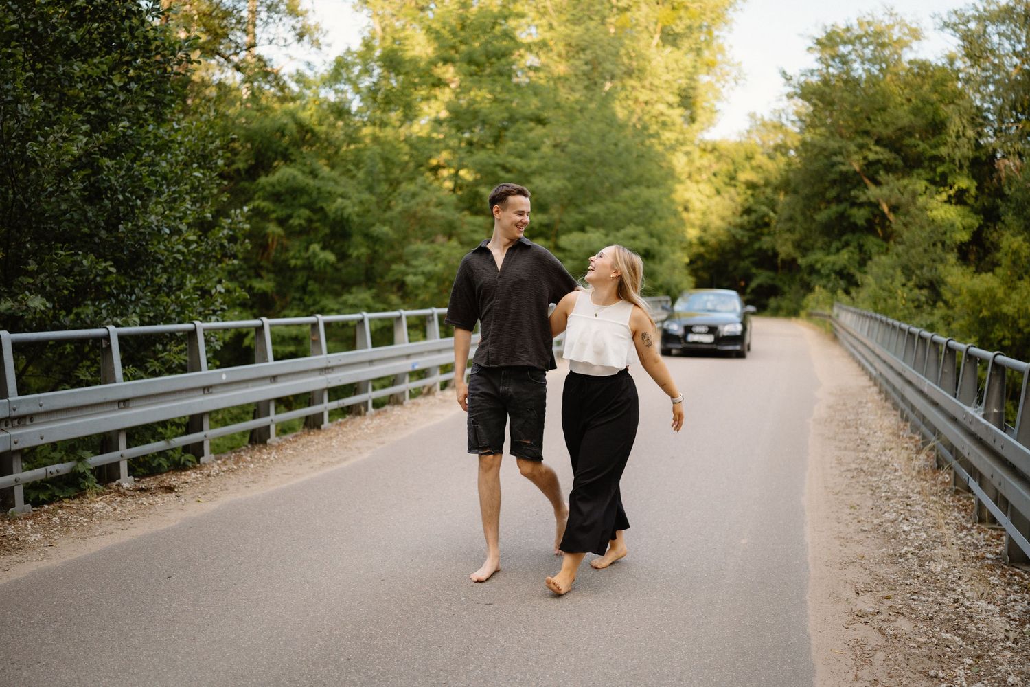 A couple walks barefoot together on a peaceful tree-lined road at sunset.