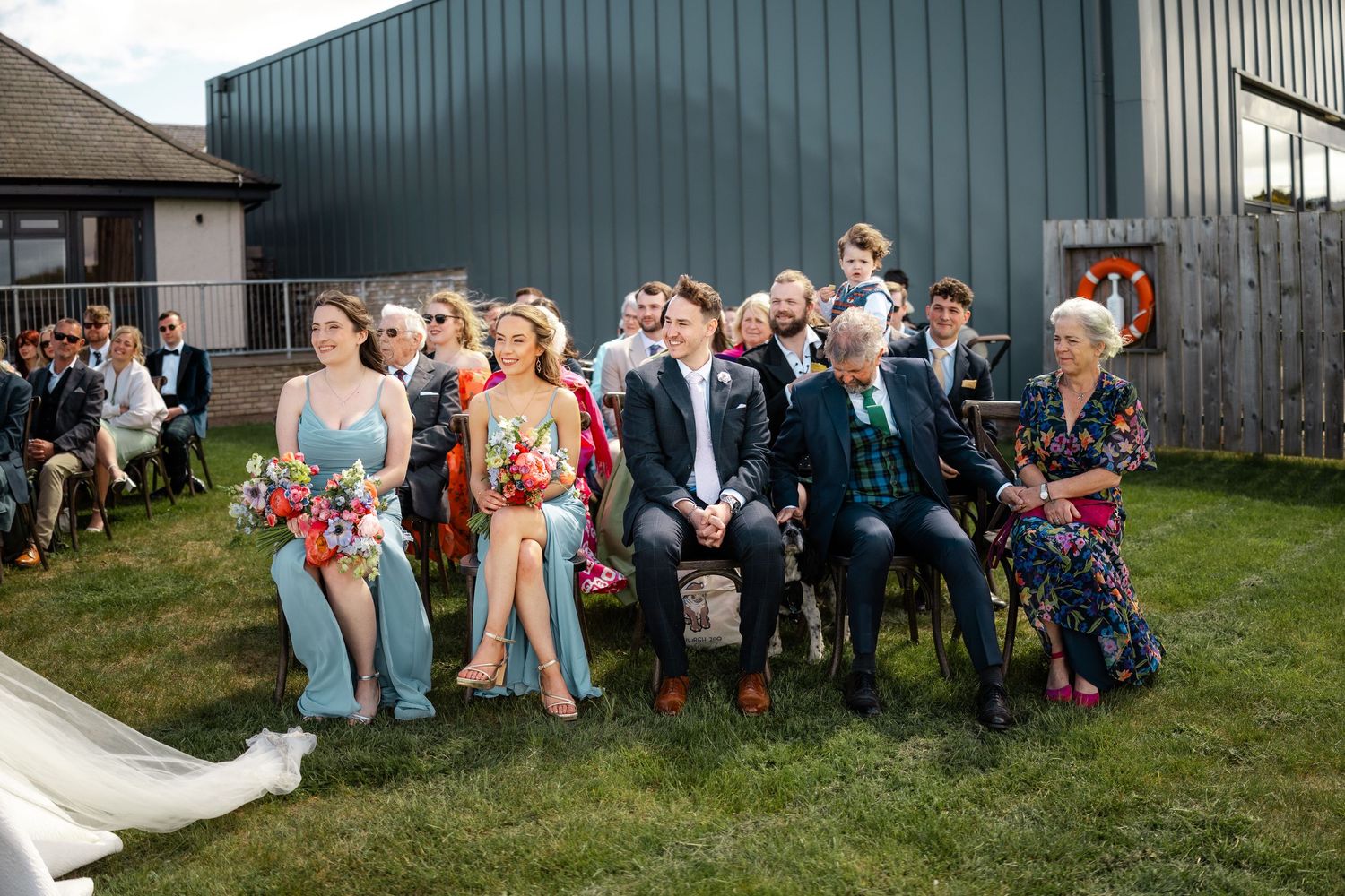 Wedding guests seated in rows on a lawn in front of a dark green building.