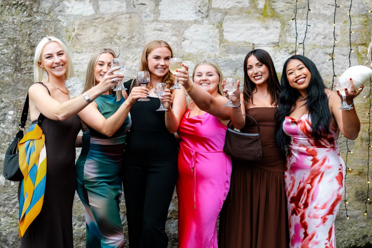 Group of wedding guests in colorful dresses pose together against stone wall.