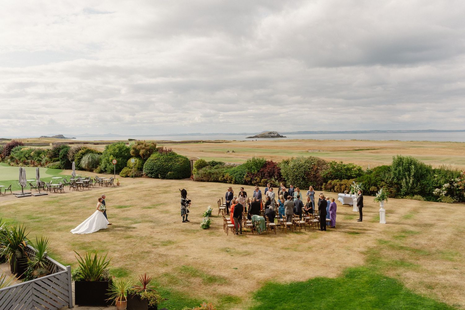 Aerial view of an outdoor wedding ceremony on a coastal lawn with guests seated facing the ocean under cloudy skies.