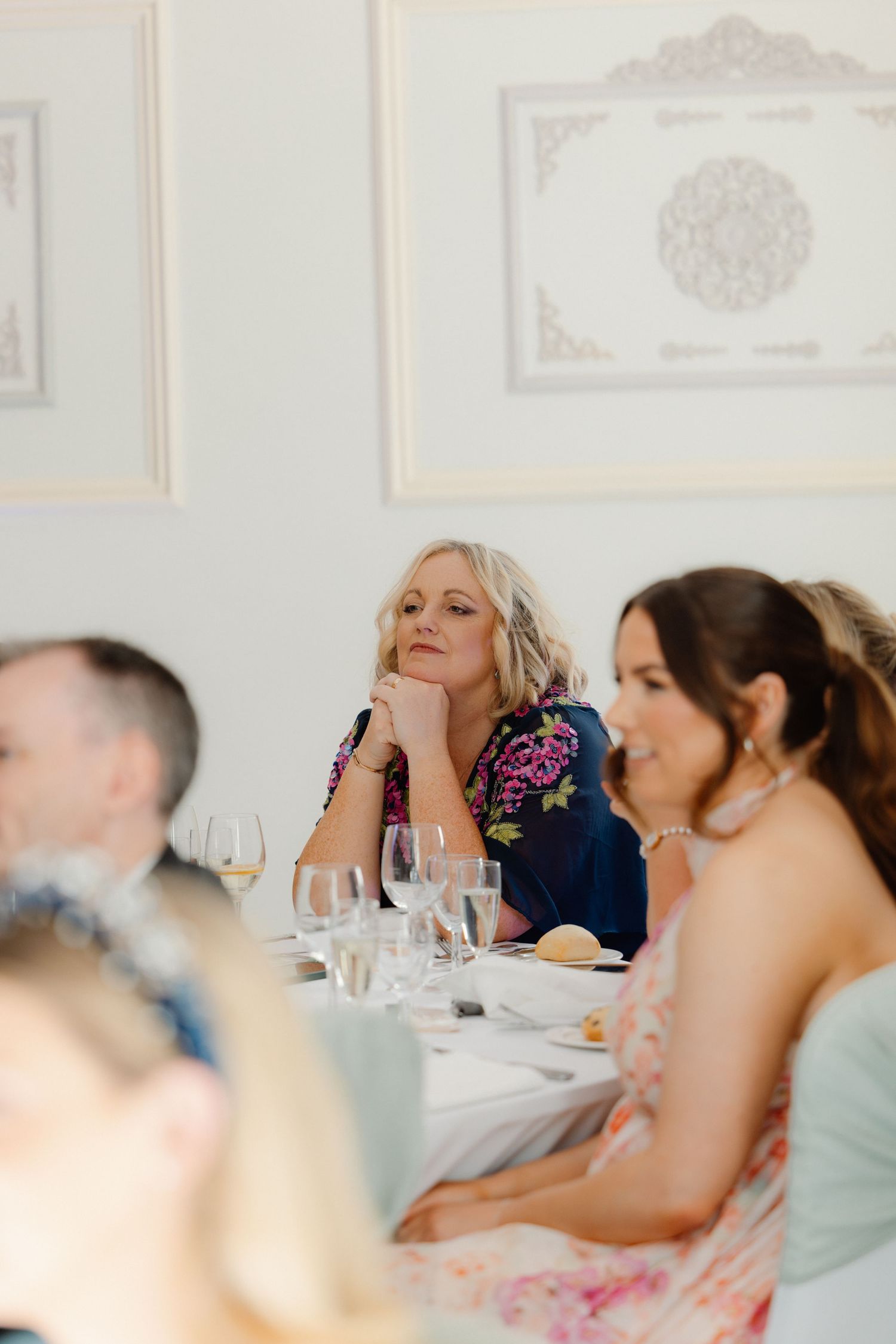Elegant wedding guest in floral dress listens attentively during reception speeches at a beautifully set dining table.