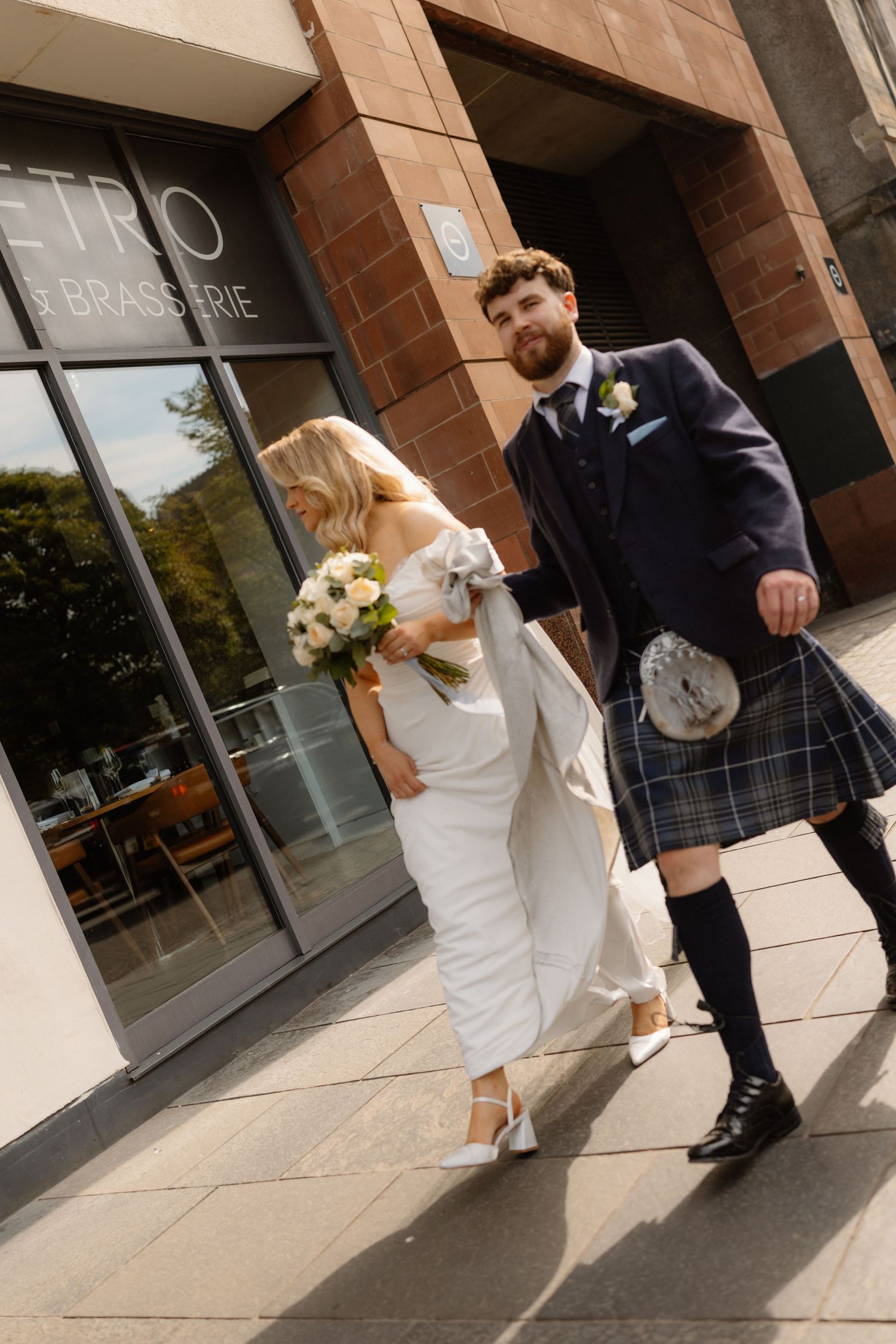 A bride in white dress and groom in Scottish kilt walk together outside a brasserie on their wedding day.