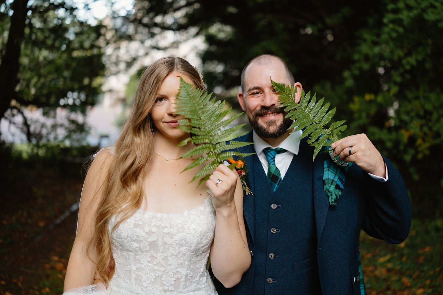 Bride and groom playfully hide behind large green fern leaves in an outdoor portrait.