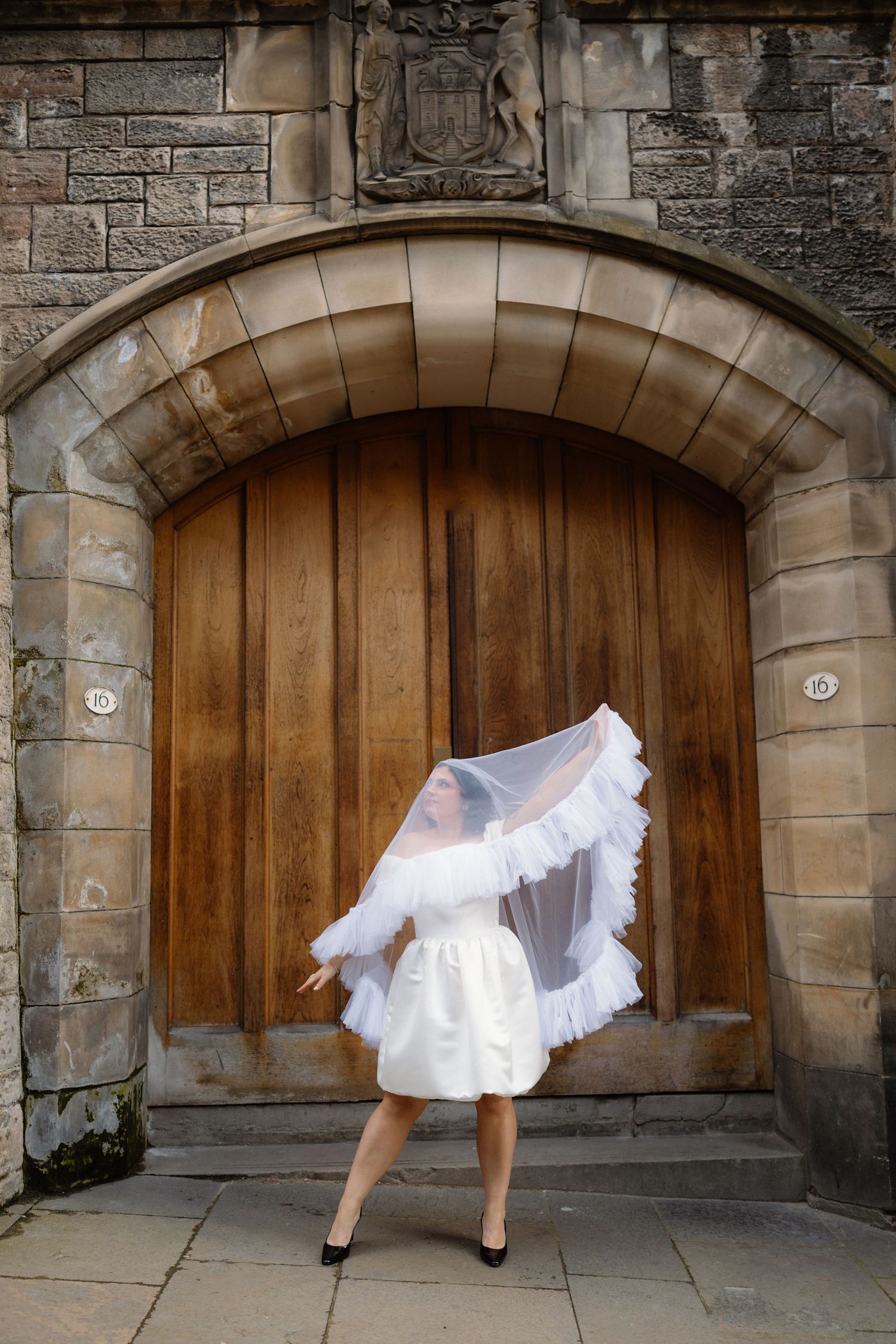 Bride in short white dress with flowing veil standing before a historic stone arched doorway.