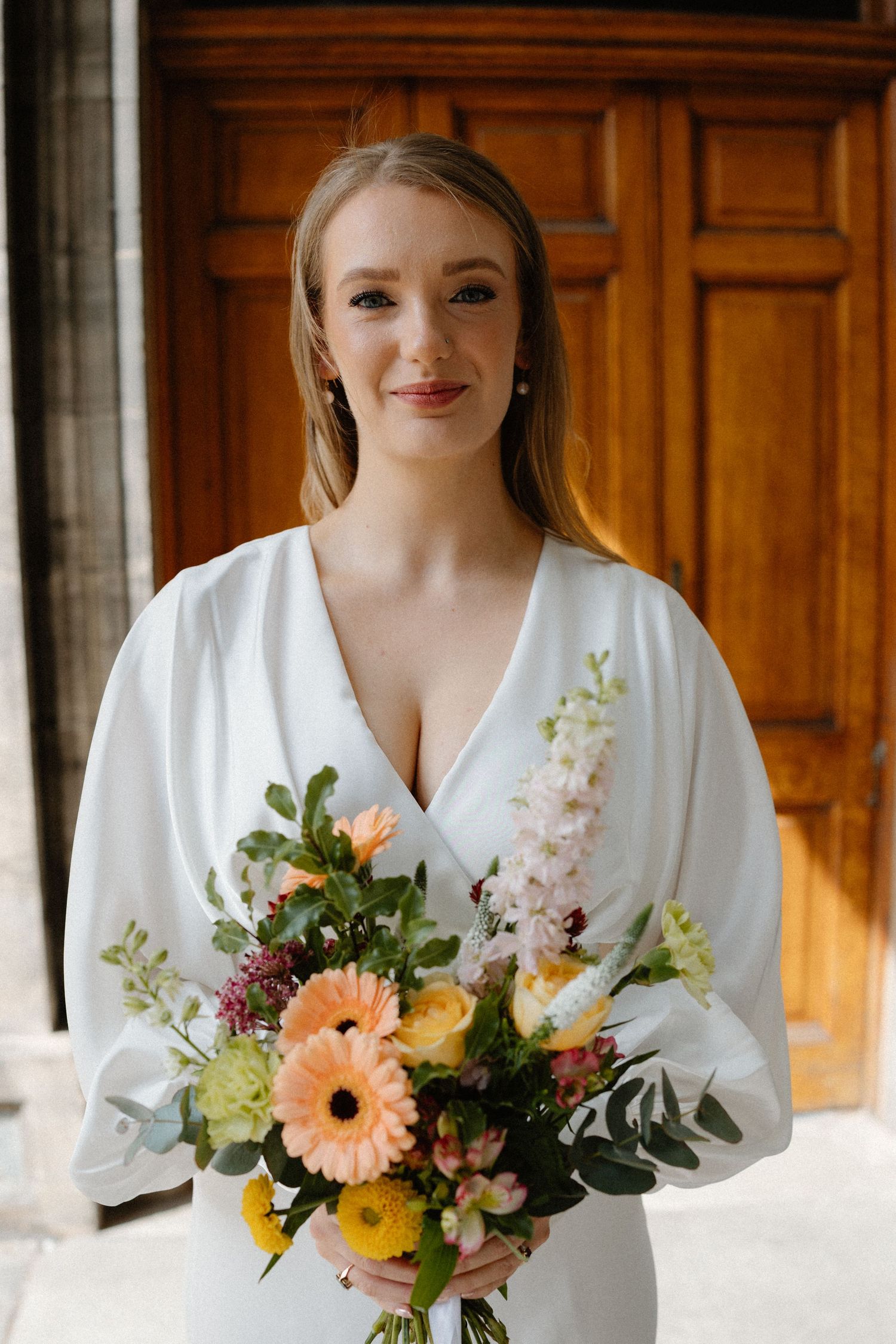 Smiling bride in white dress holds colorful bouquet with gerberas and wildflowers before wooden doors.