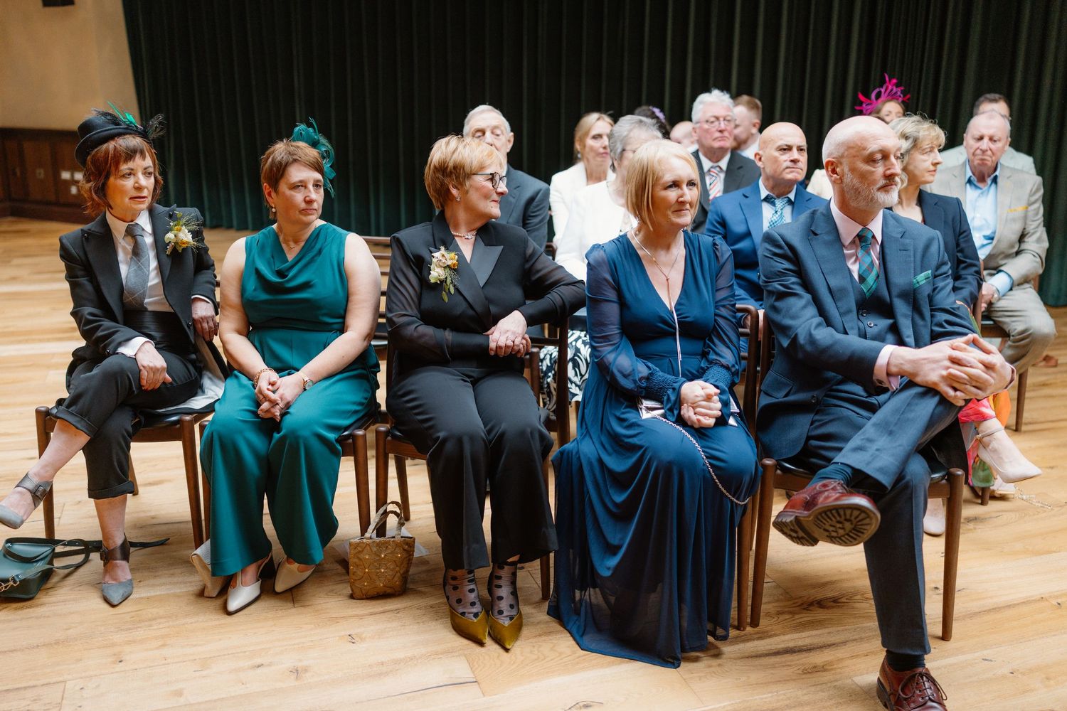 Elegantly dressed wedding guests seated in rows at a ceremony, wearing teal and green formal attire in a wood-floored hall.