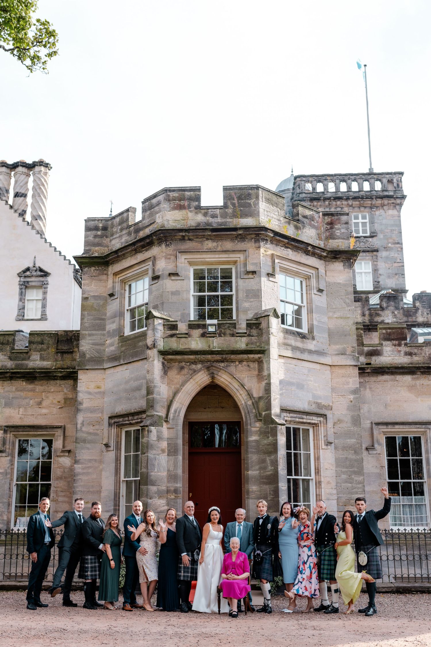 Wedding party and guests pose together outside a historic Scottish stone castle venue under a cloudy sky.