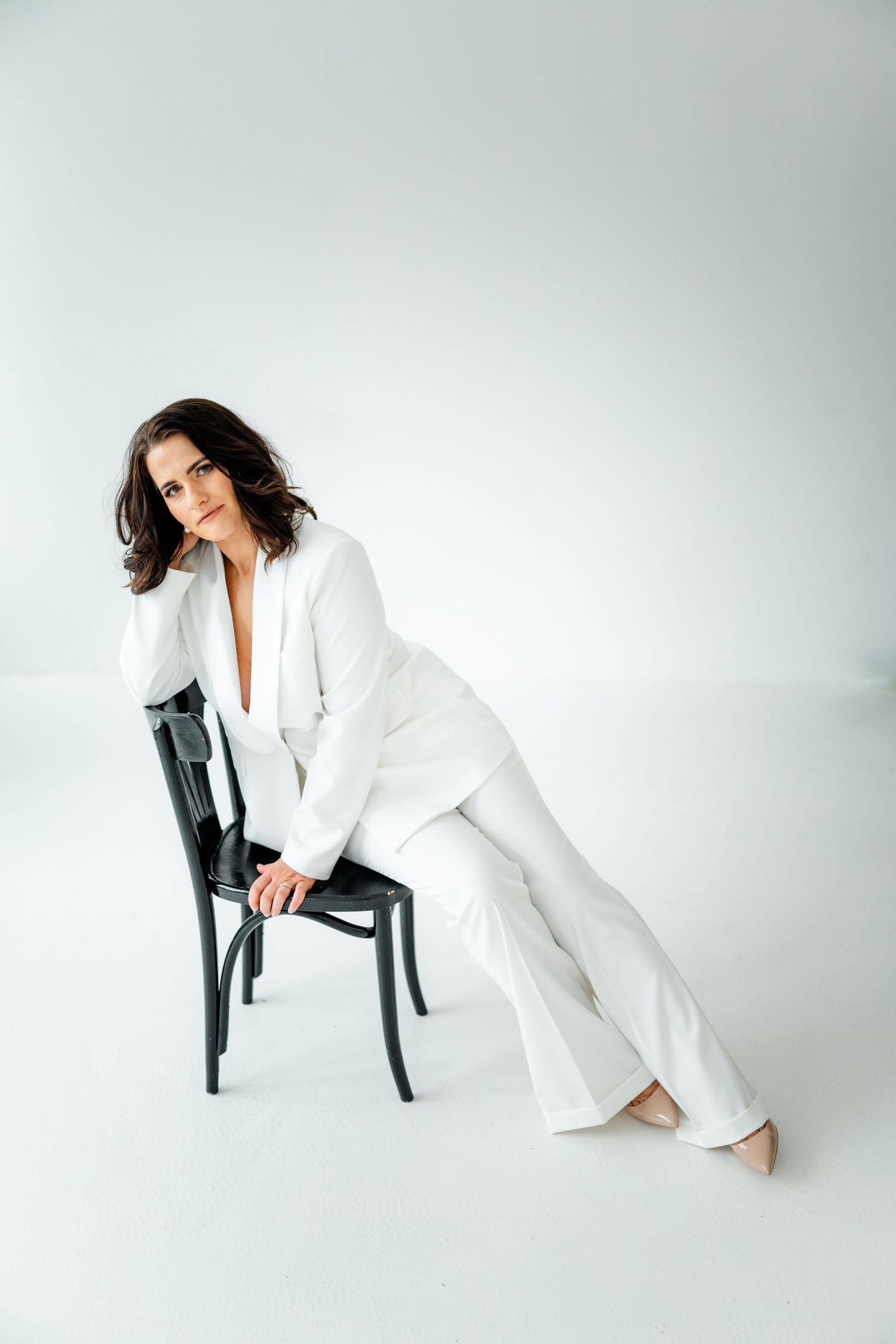Model in white suit poses elegantly with modern black chair in studio setting.