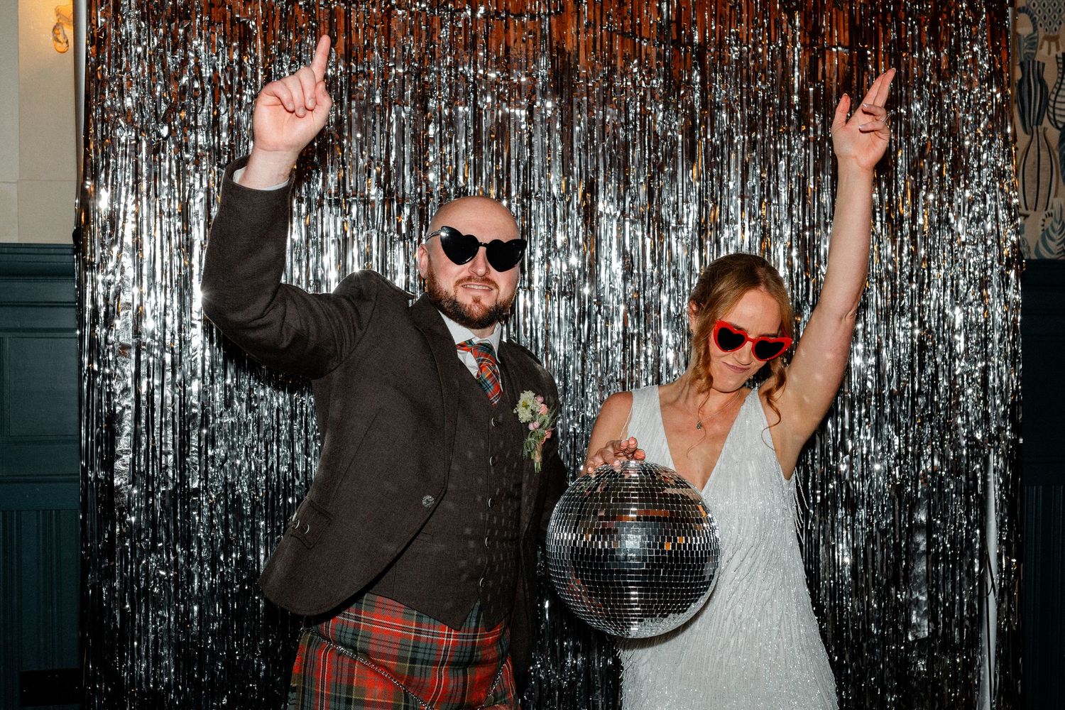 A couple celebrates with raised arms in front of a silver tinsel backdrop.