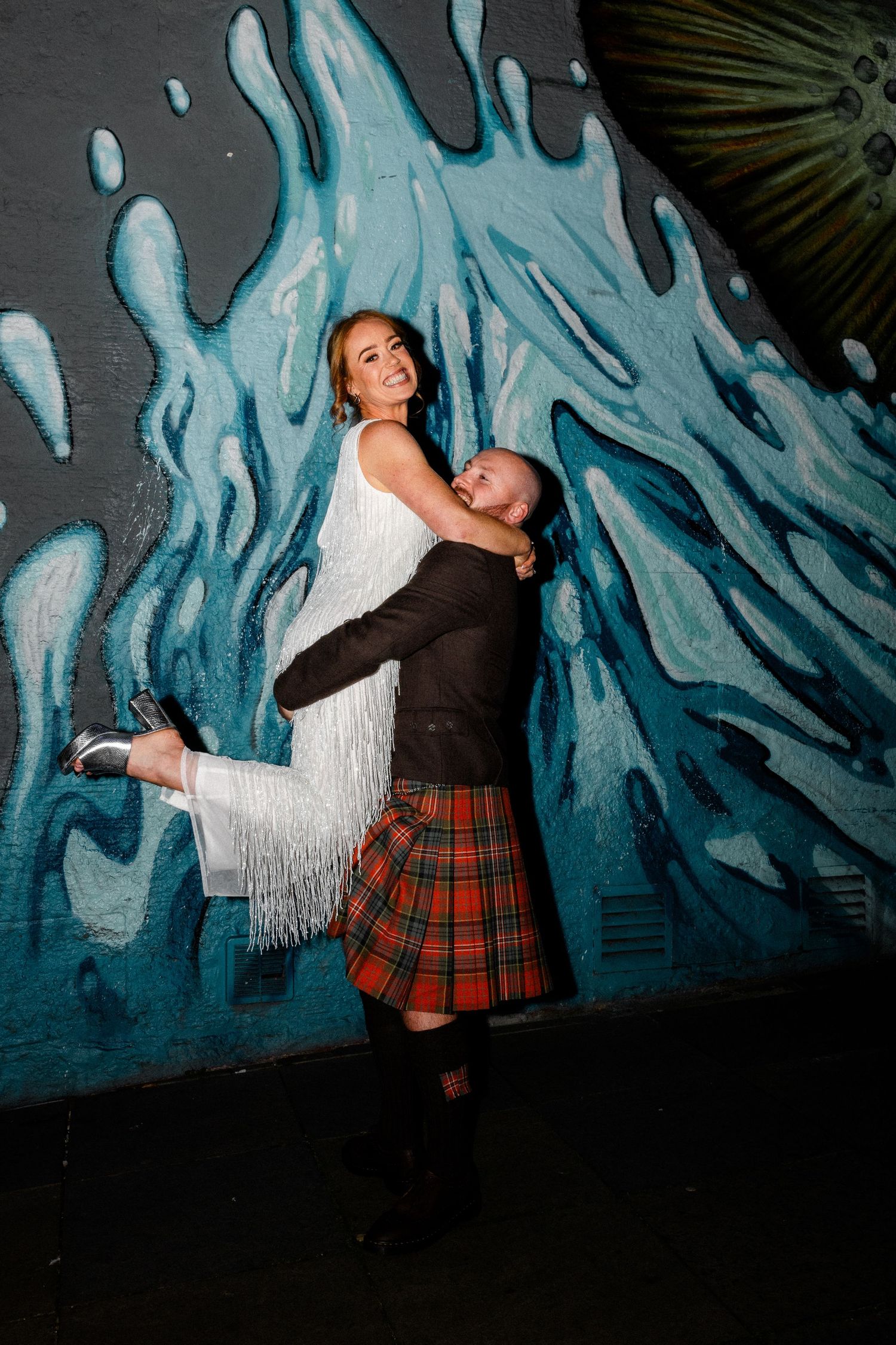 A couple posing playfully against a blue painted wall mural at night.