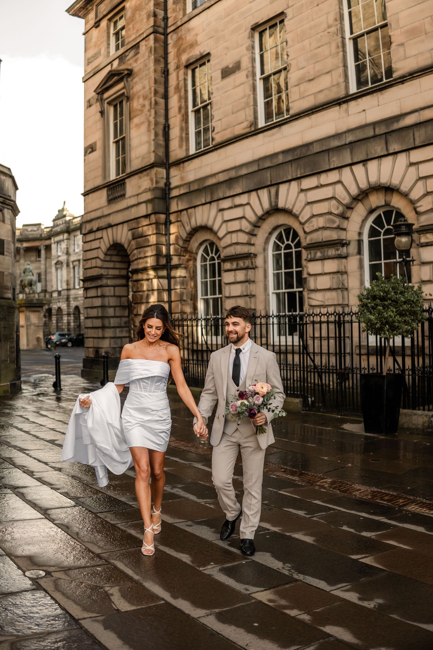 Couple strolling on wet cobblestone street beside historic Edinburgh building.