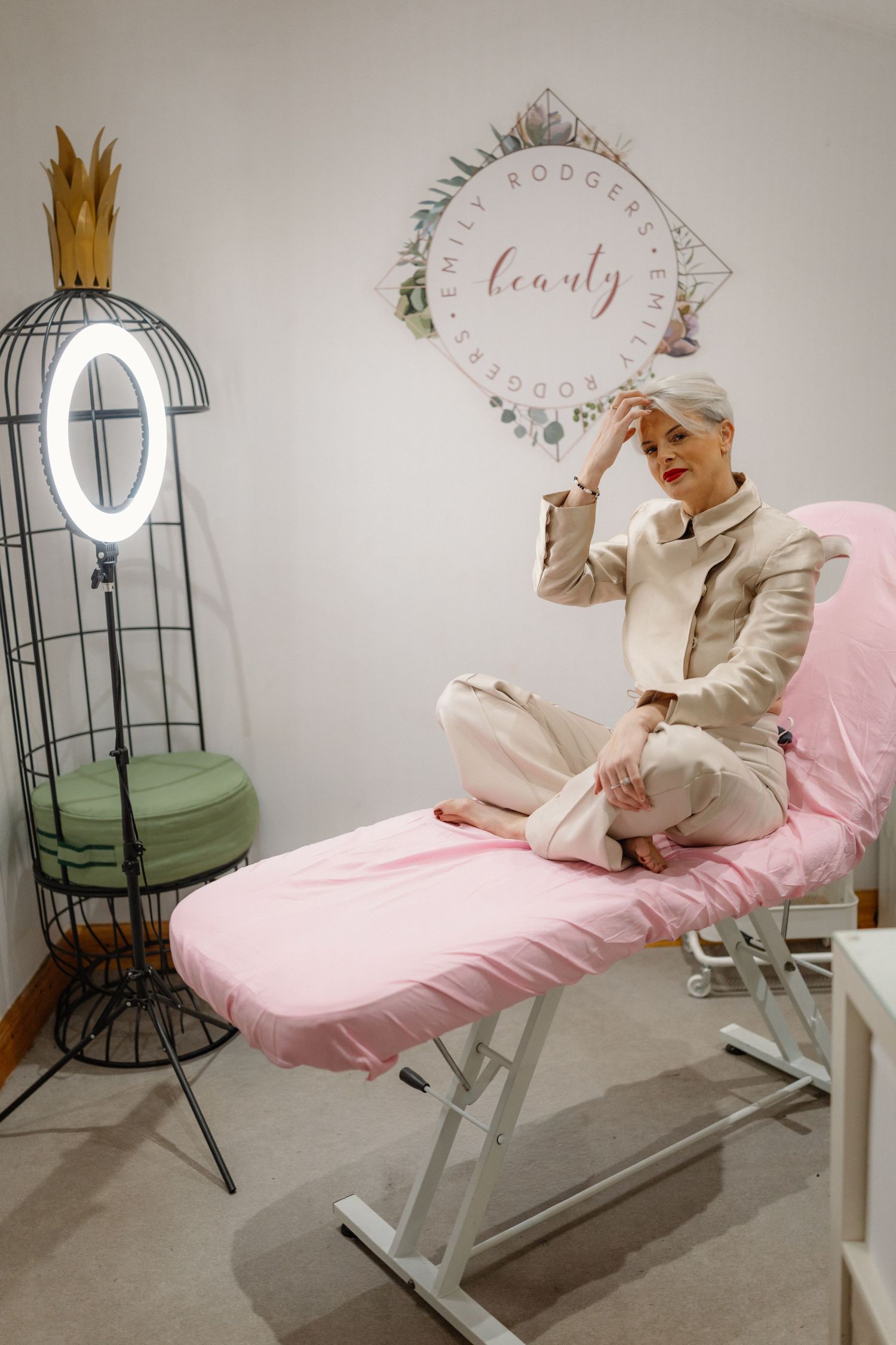 A person in white clothing and crown poses on a pink reclining chair in a minimalist room with decorative elements.