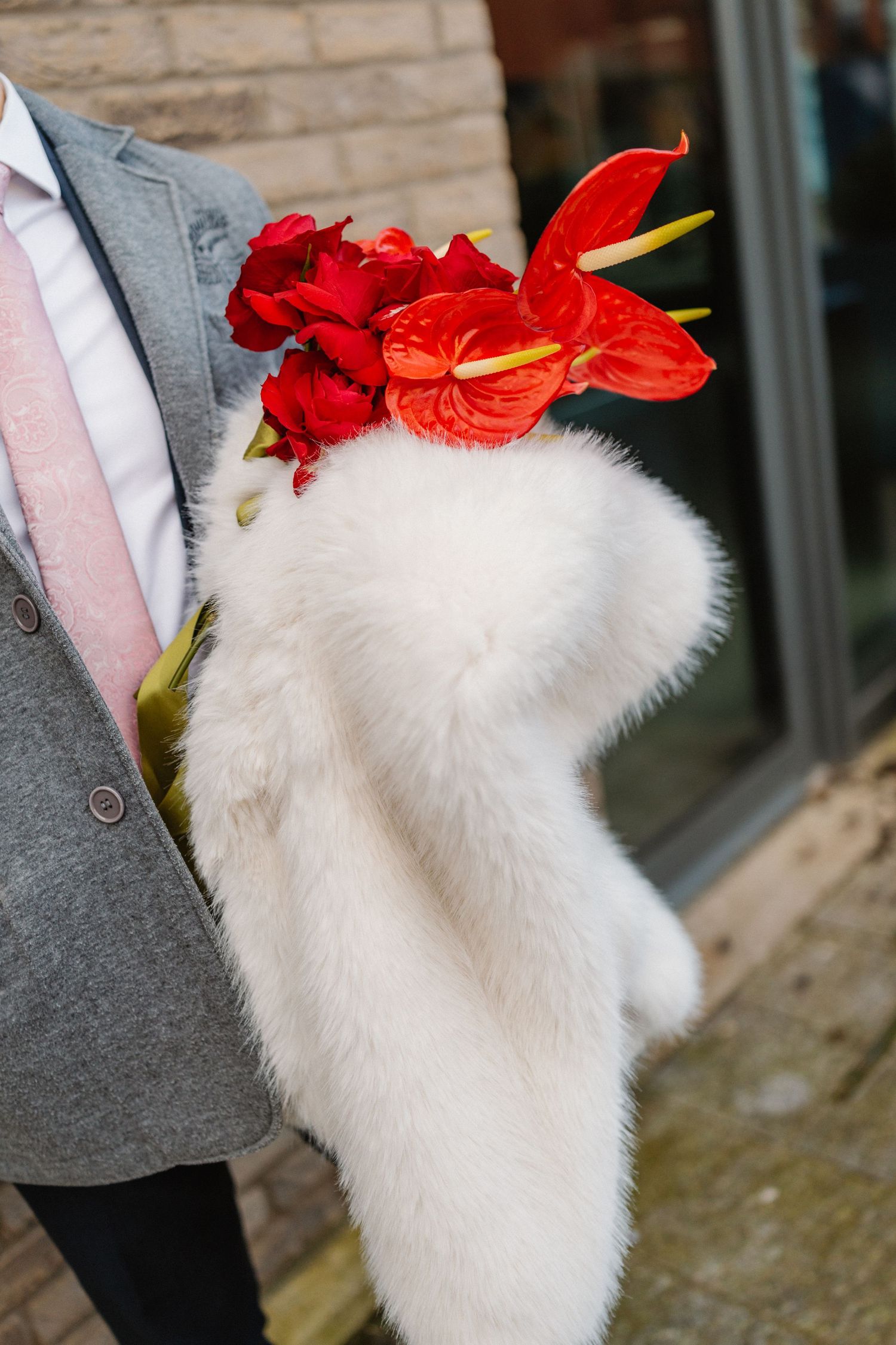 Groom in gray suit holding white fur muff bouquet with red anthuriums and roses at wedding.