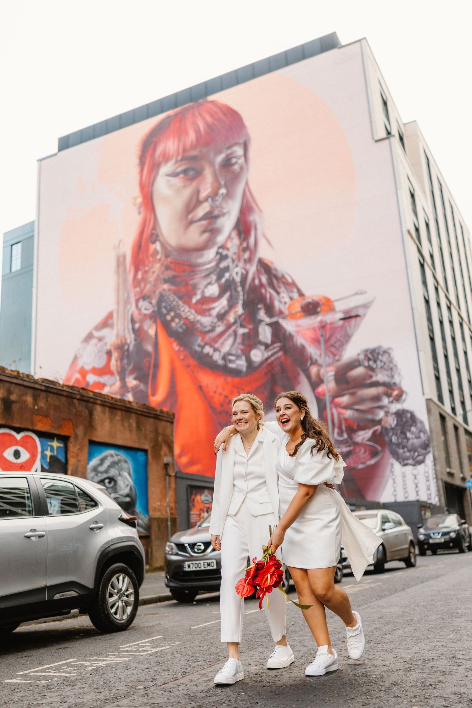 Two brides in white outfits laughing and walking past a large street mural in an urban setting.