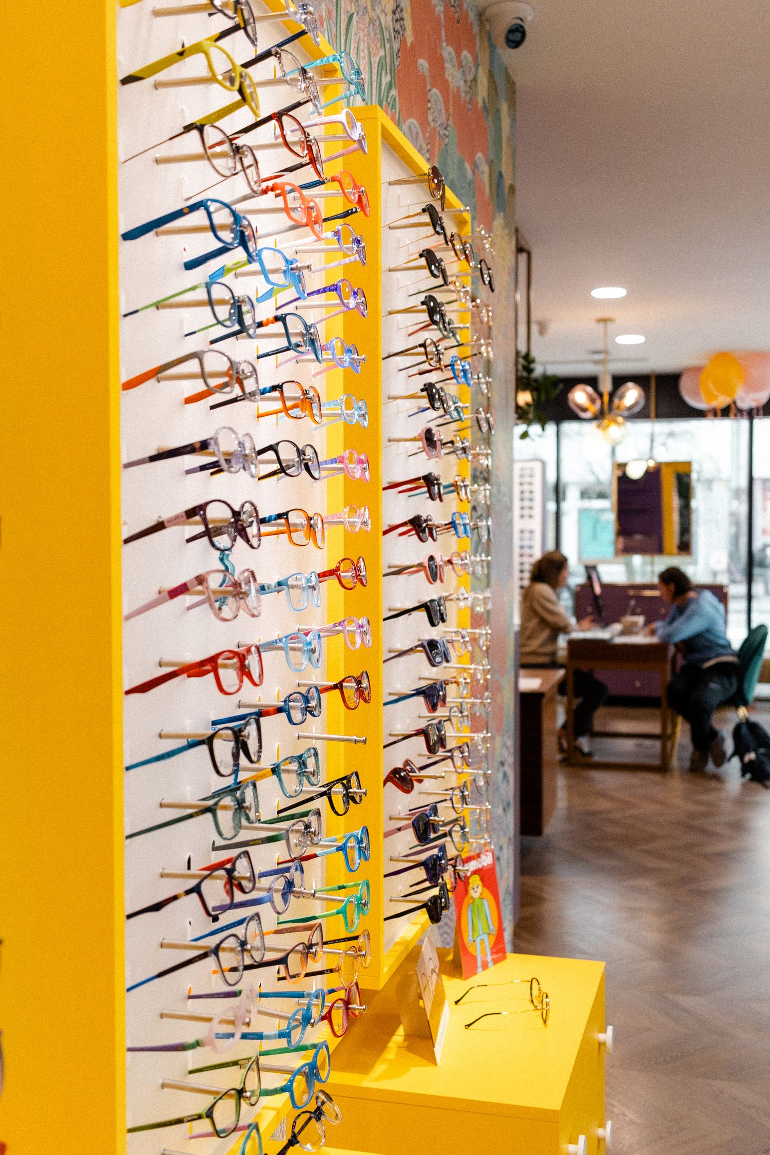 Colorful display of eyeglass frames mounted on bright yellow wall in optical store.