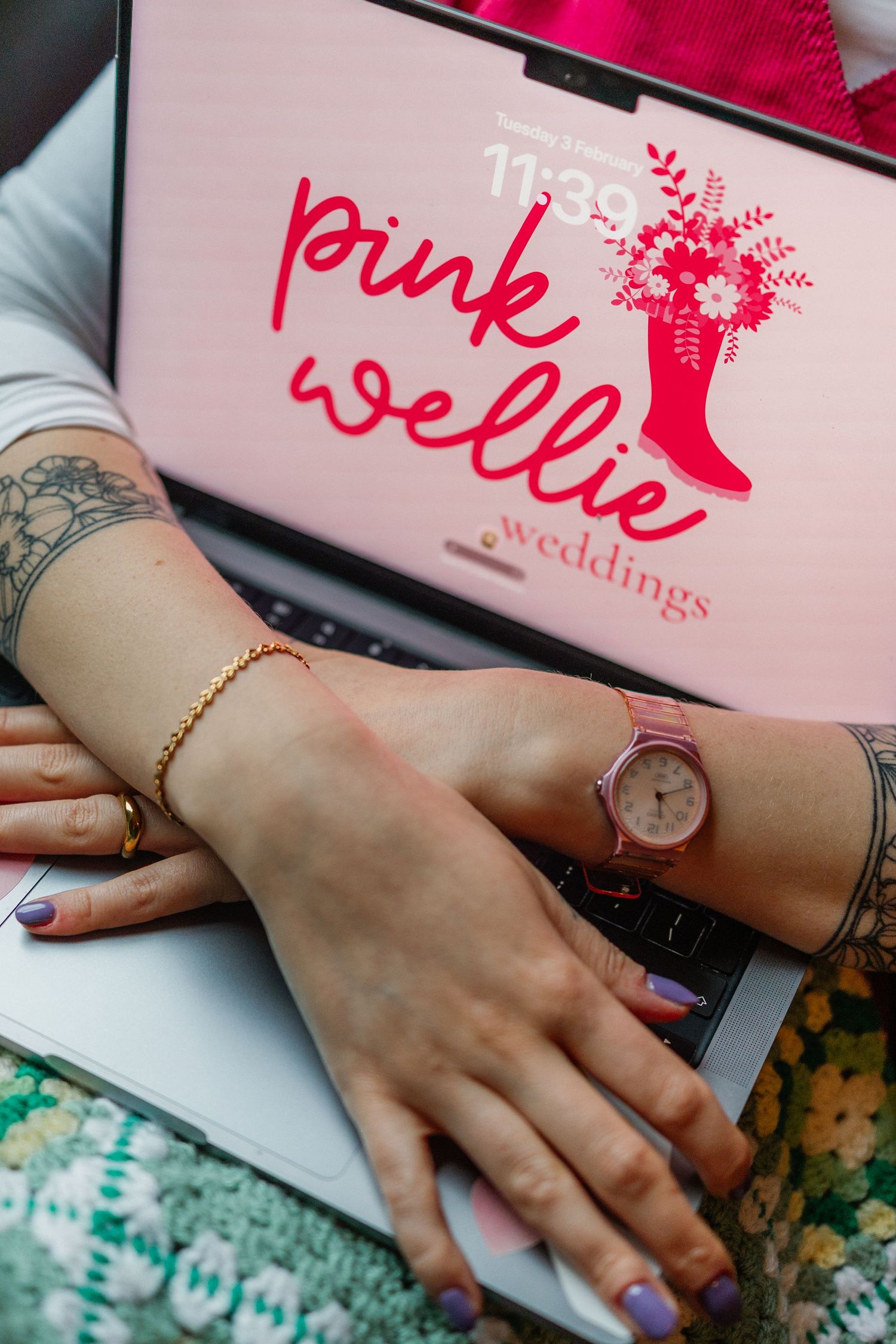 Close-up of hands wearing a gold bracelet and watch next to pink wedding branding materials.