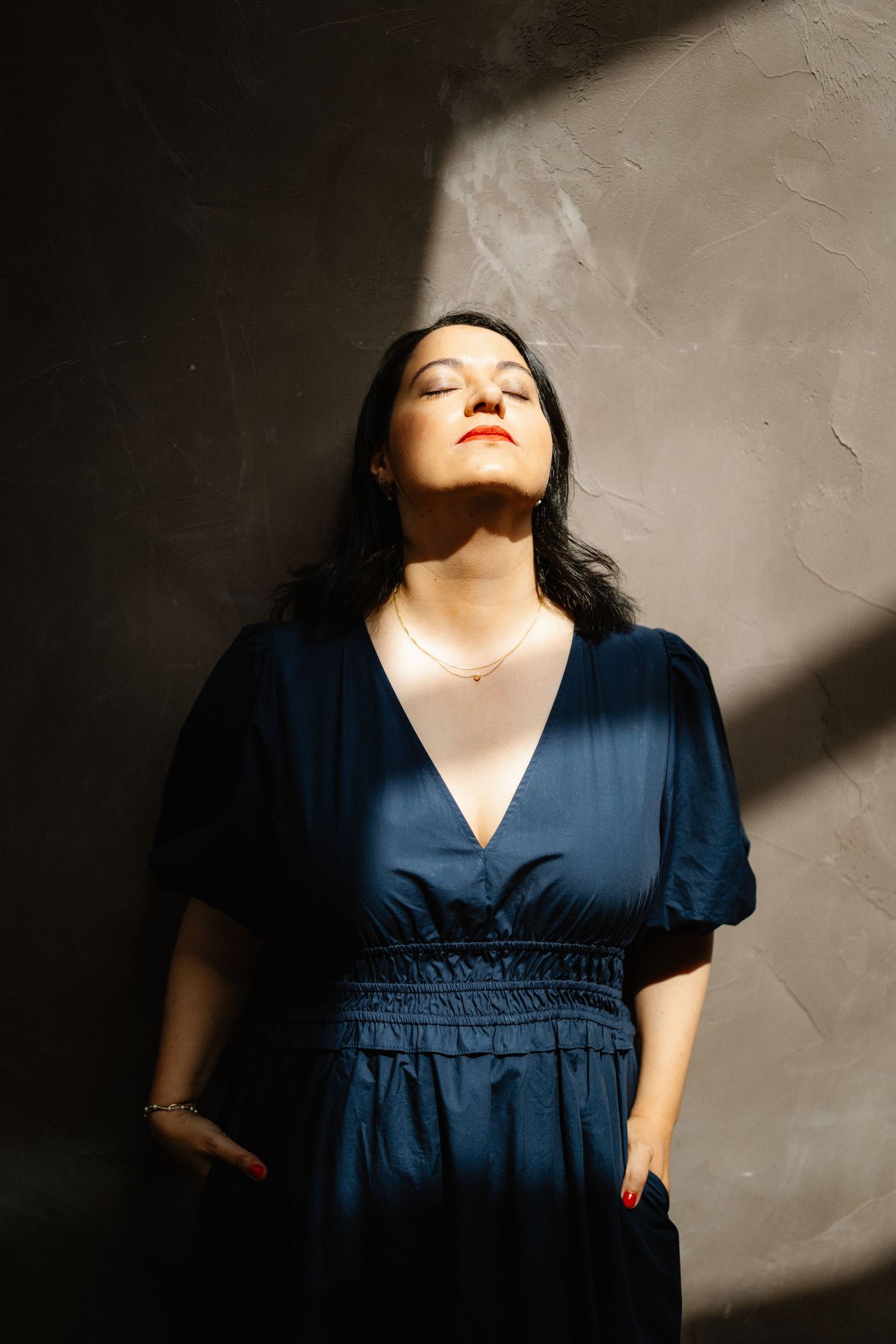 Woman in navy blue dress poses against textured wall with dramatic lighting and shadows.