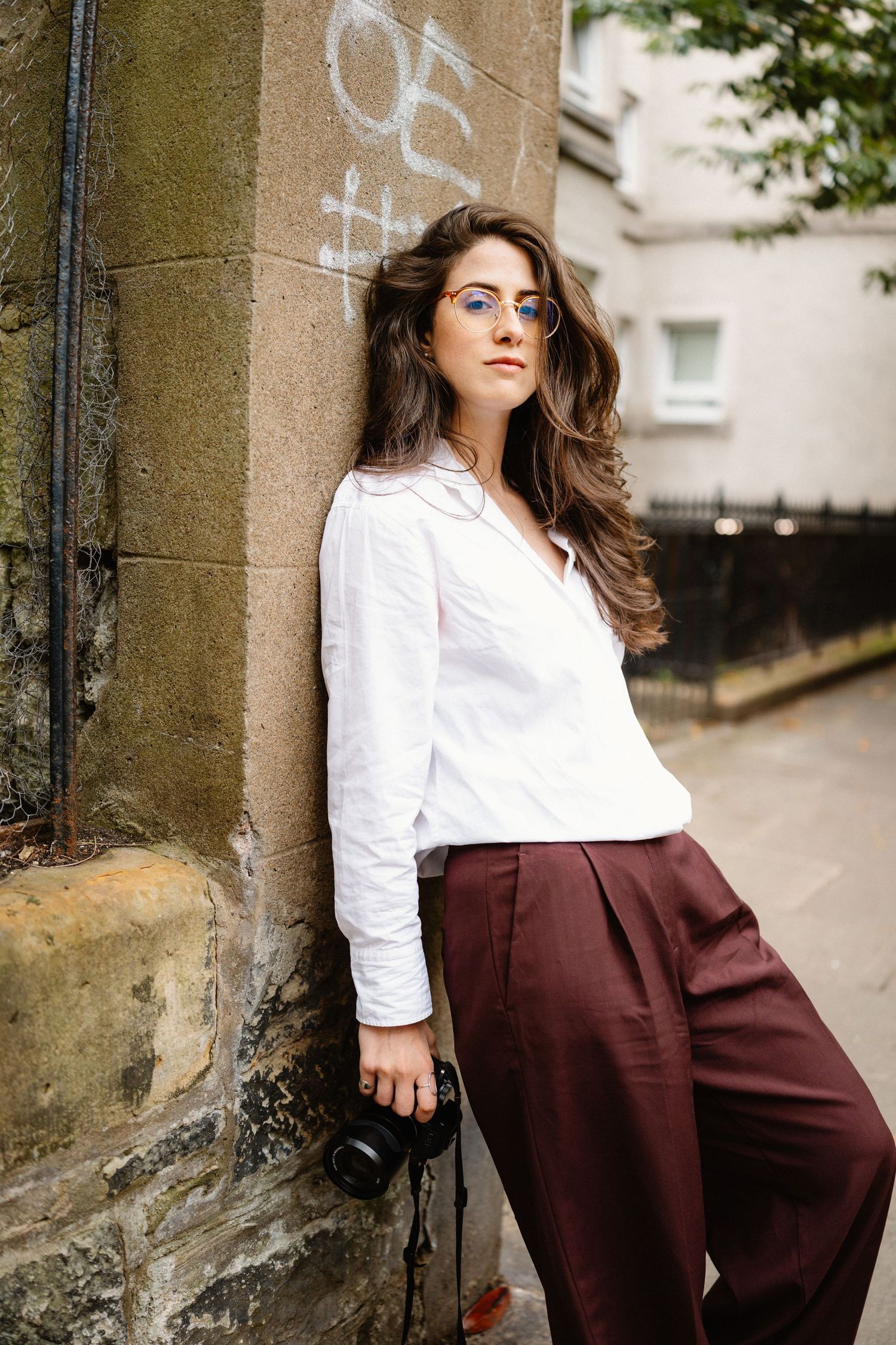 Fashion model poses in white blouse and burgundy pants against stone wall.