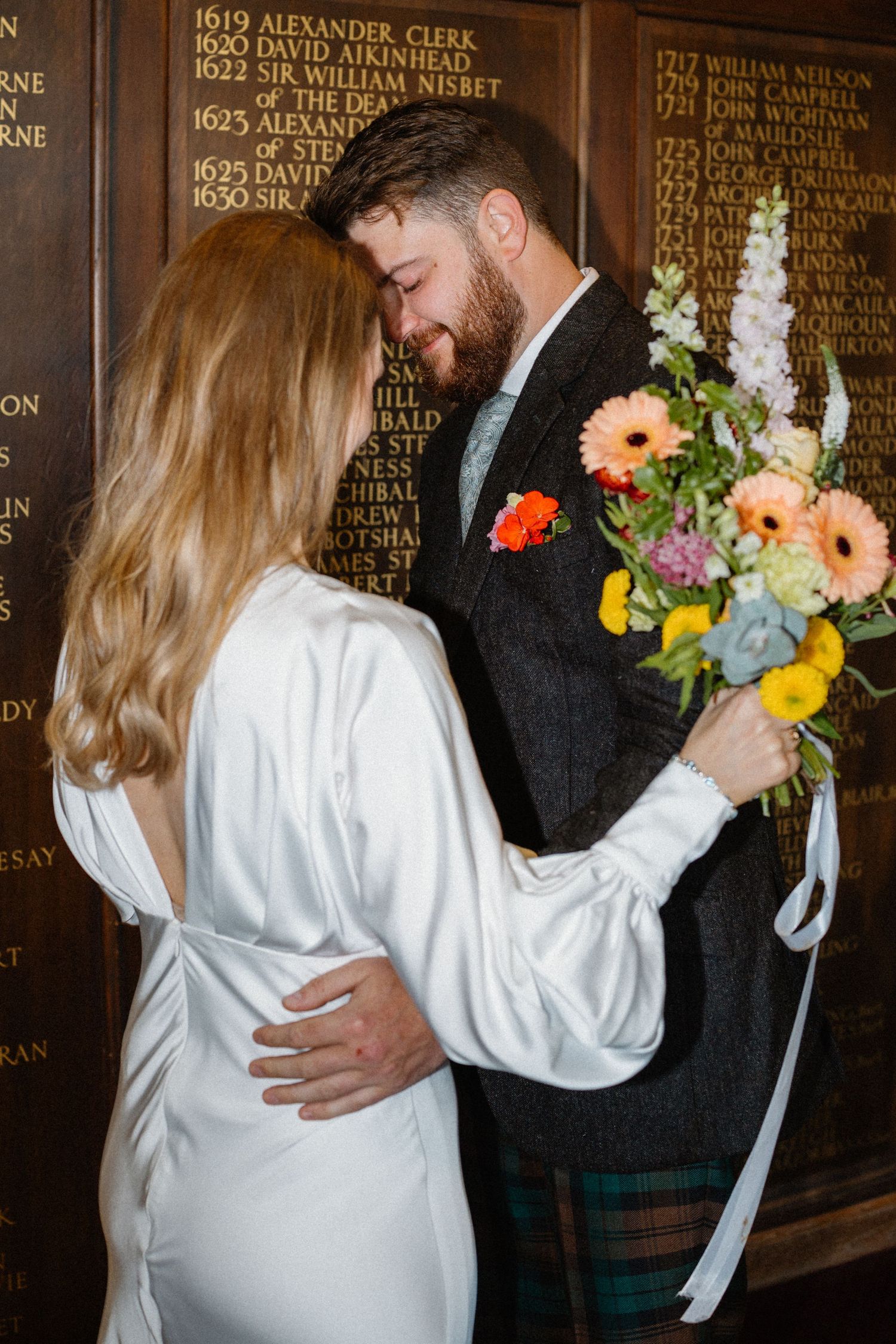 A couple in wedding attire embraces while holding a colorful bouquet of flowers against a wall with engraved names.