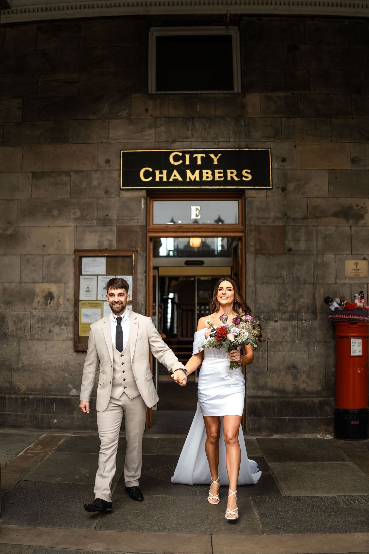 A couple exits City Chambers building after their wedding ceremony, with a red postbox visible in the background.