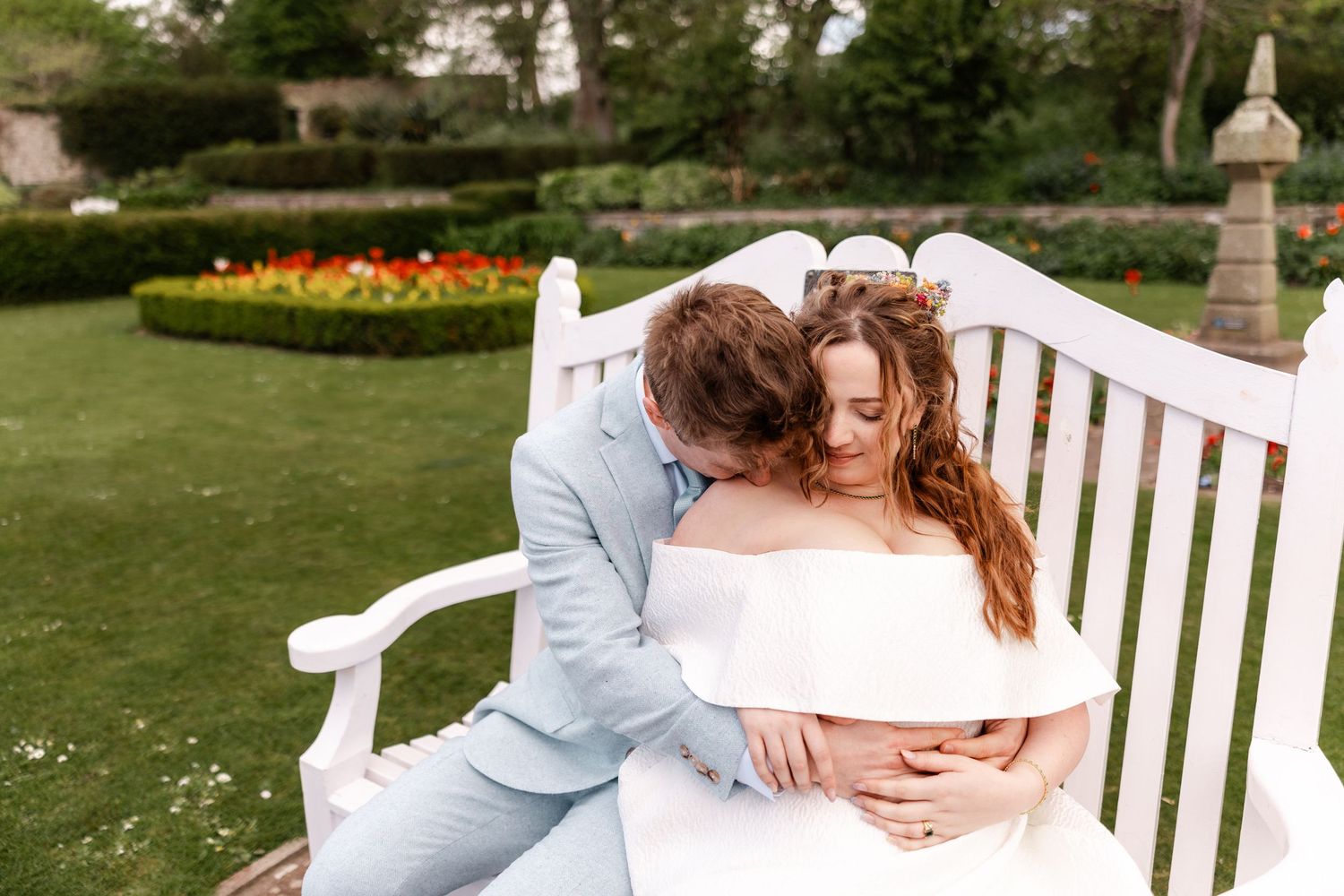 Romantic couple embracing on a white garden bench surrounded by lush greenery and flowers.