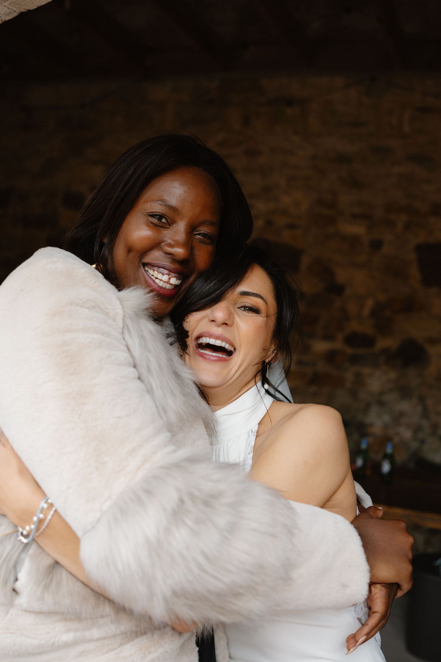 Two wedding guests share joyful embrace and laughter against rustic brick wall.