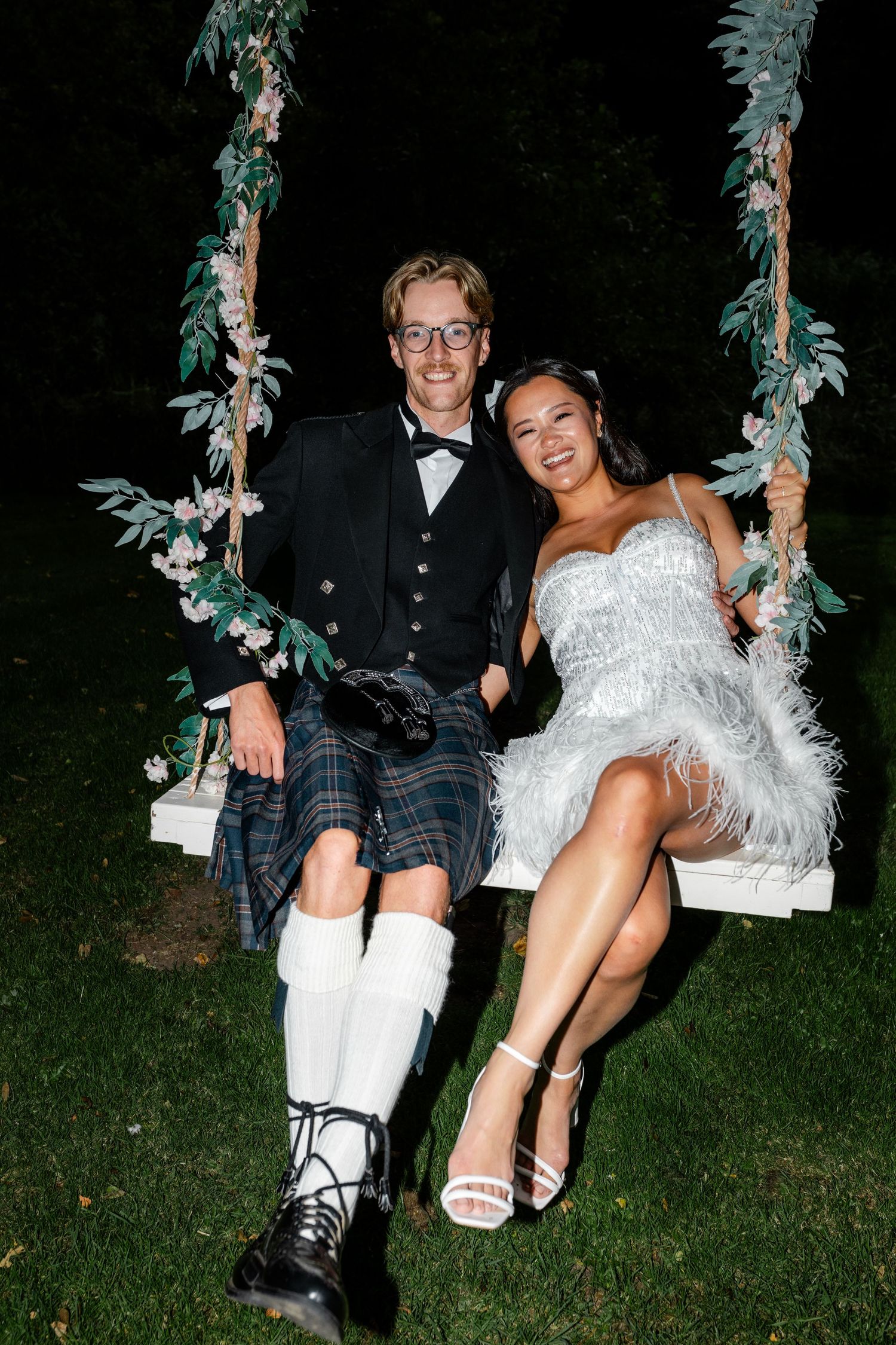 Couple sitting on floral decorated swing at outdoor wedding celebration.