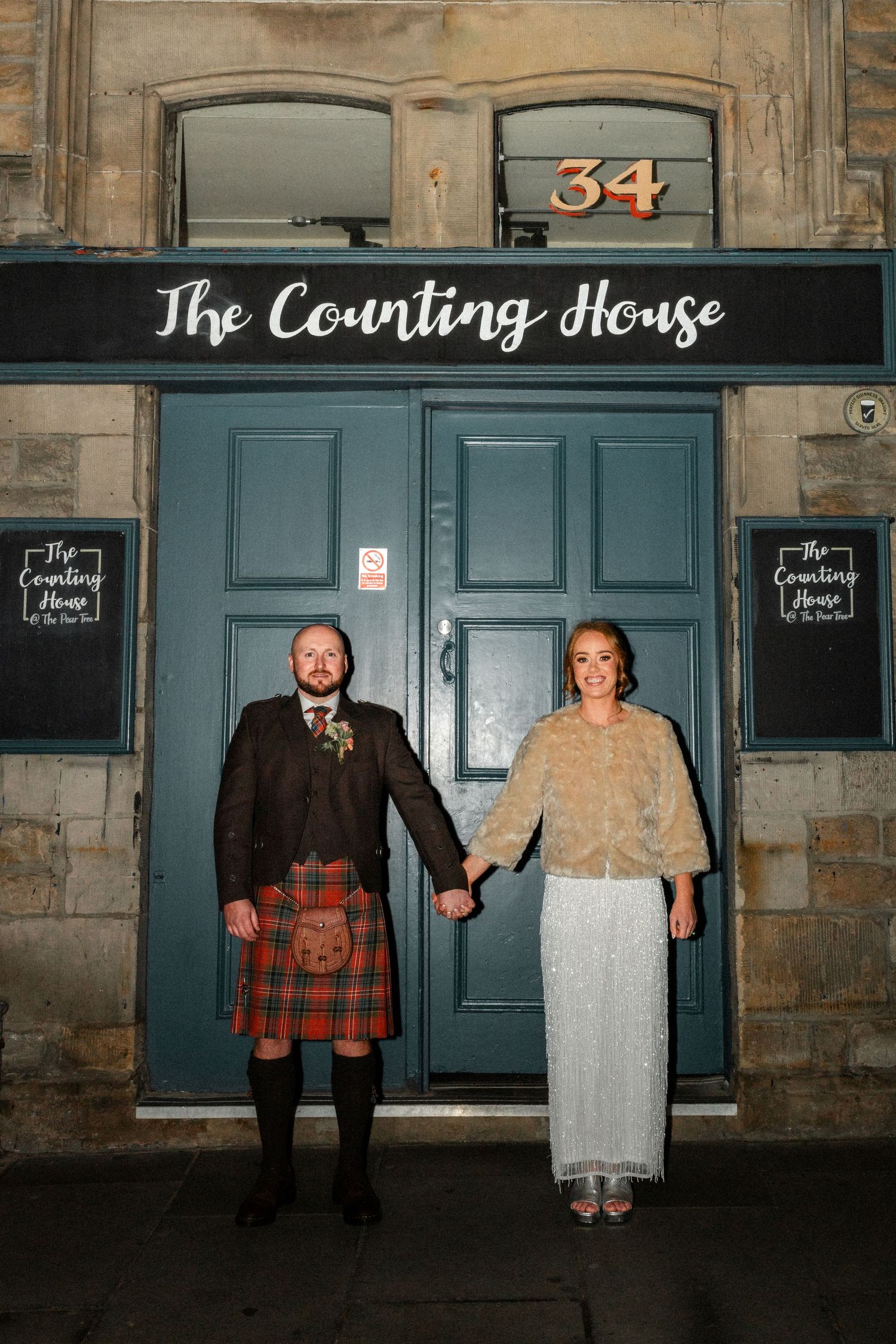 A couple poses outside The Counting House building with blue doors and stone walls at night.