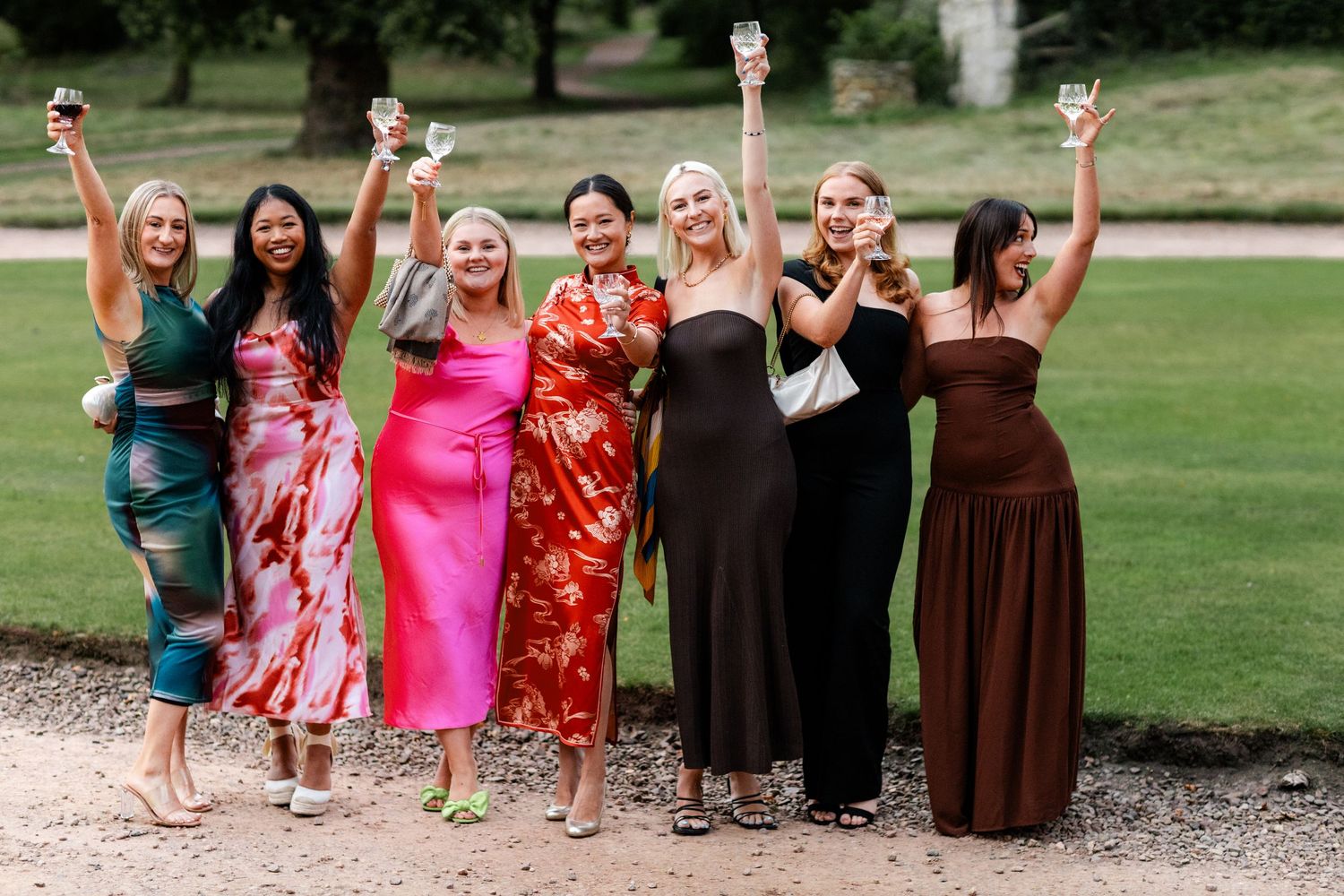 A group in vibrant dresses poses together outdoors raising their drinks in celebration.