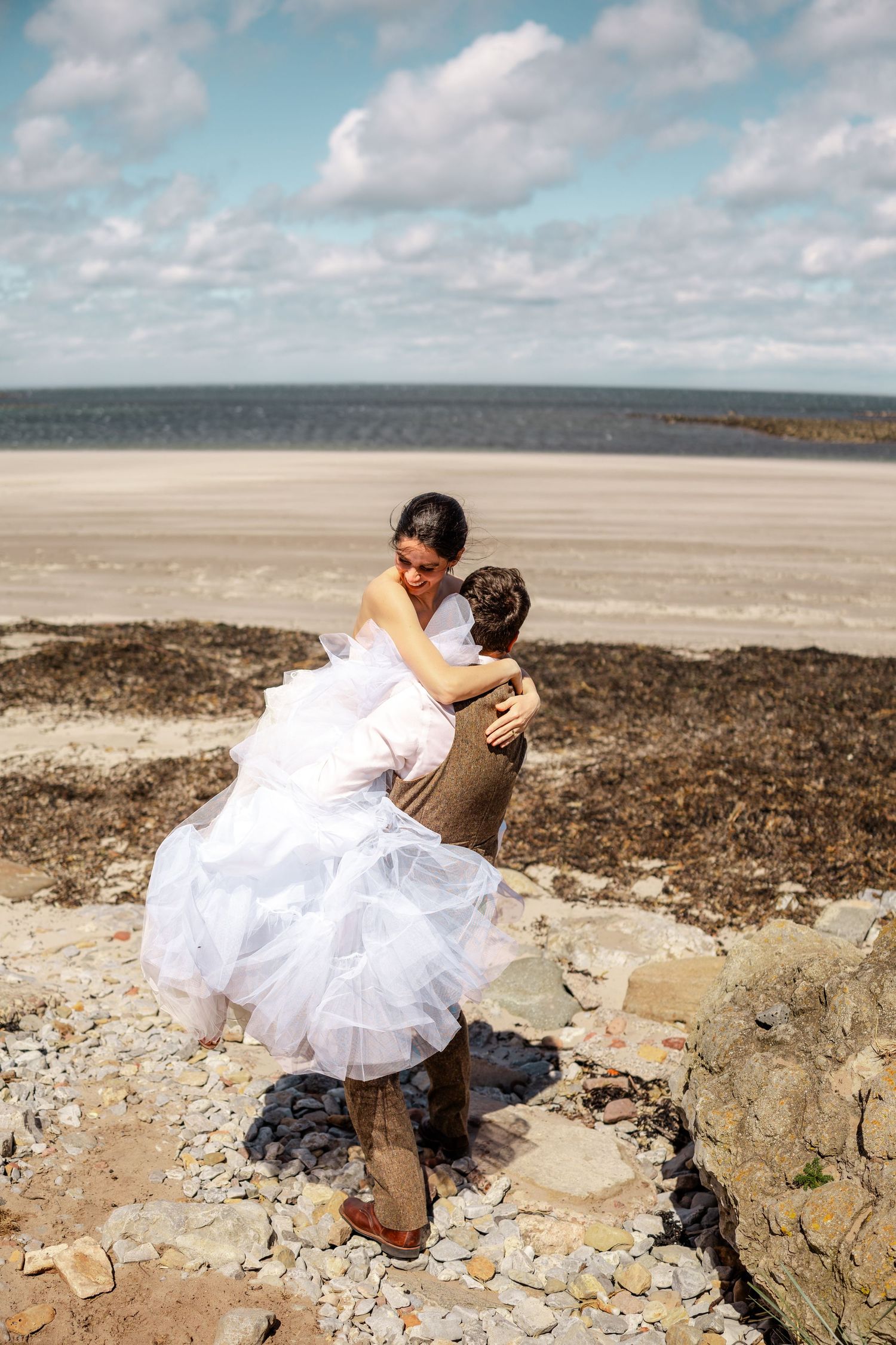 A couple in wedding attire sits on rocky coastal cliffs overlooking a scenic beach landscape.