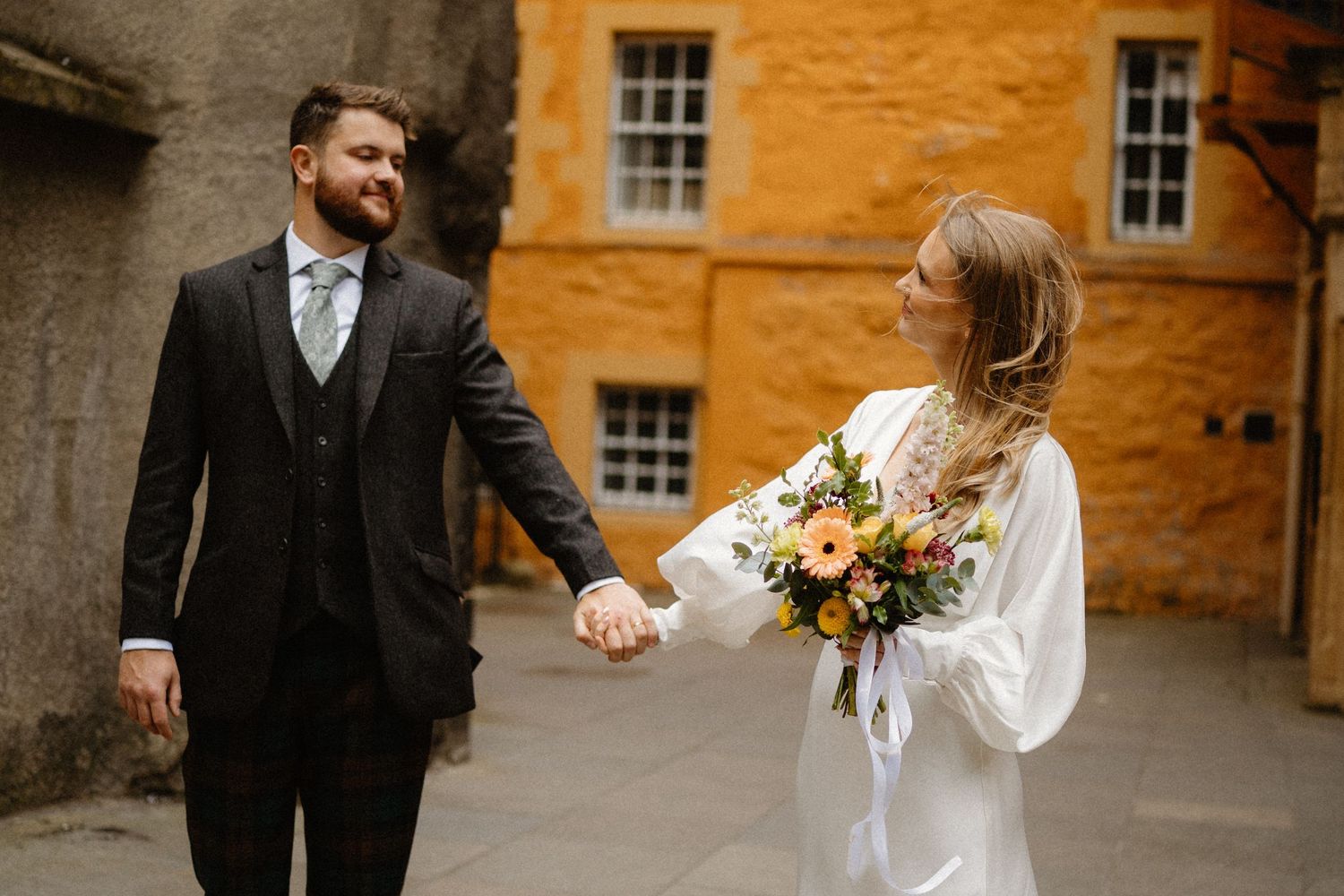 Bride and groom holding hands outdoors, bride in white dress with orange bouquet, groom in dark suit.