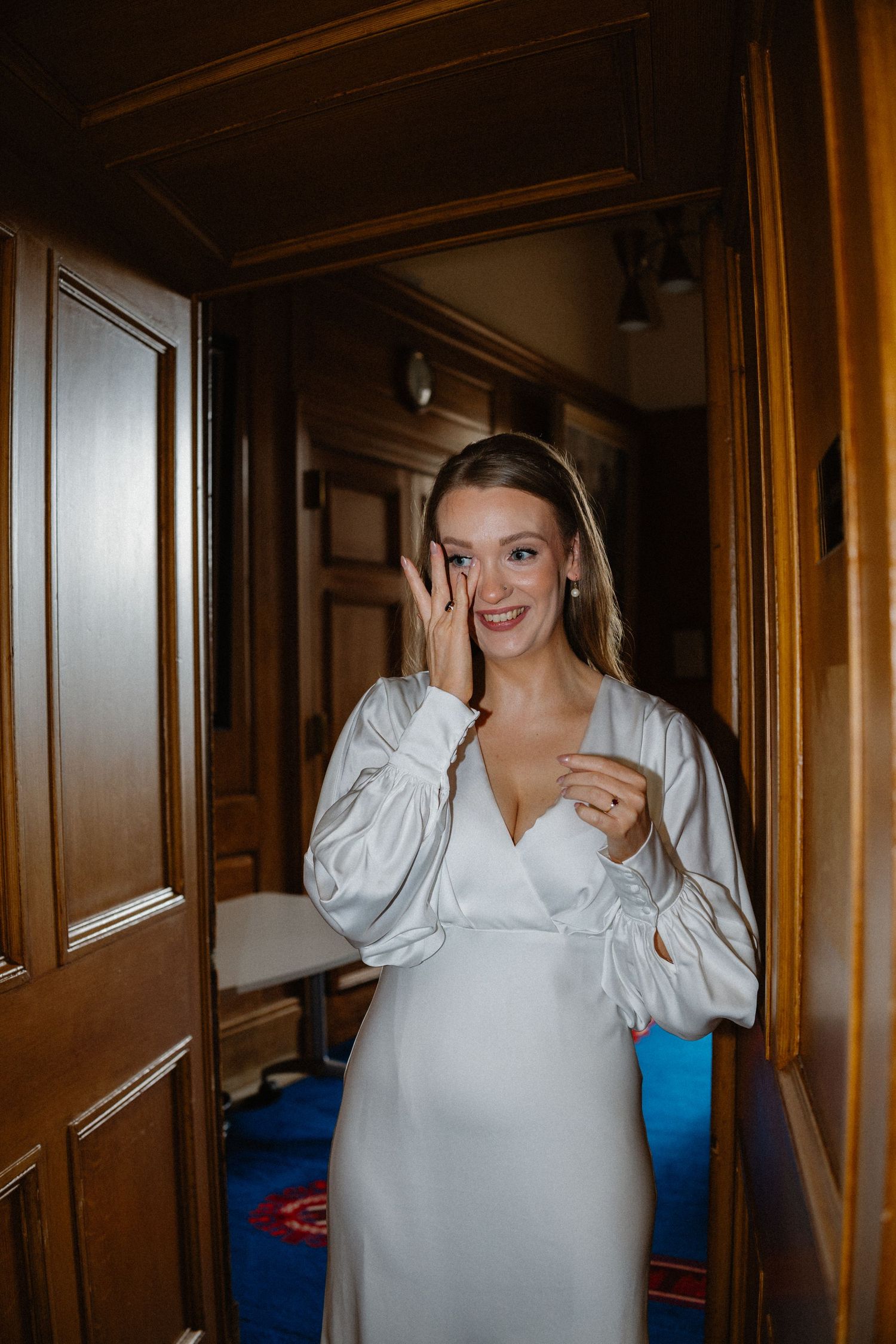 Smiling bride in elegant white satin long-sleeve wedding dress standing in wooden doorway wiping a happy tear.