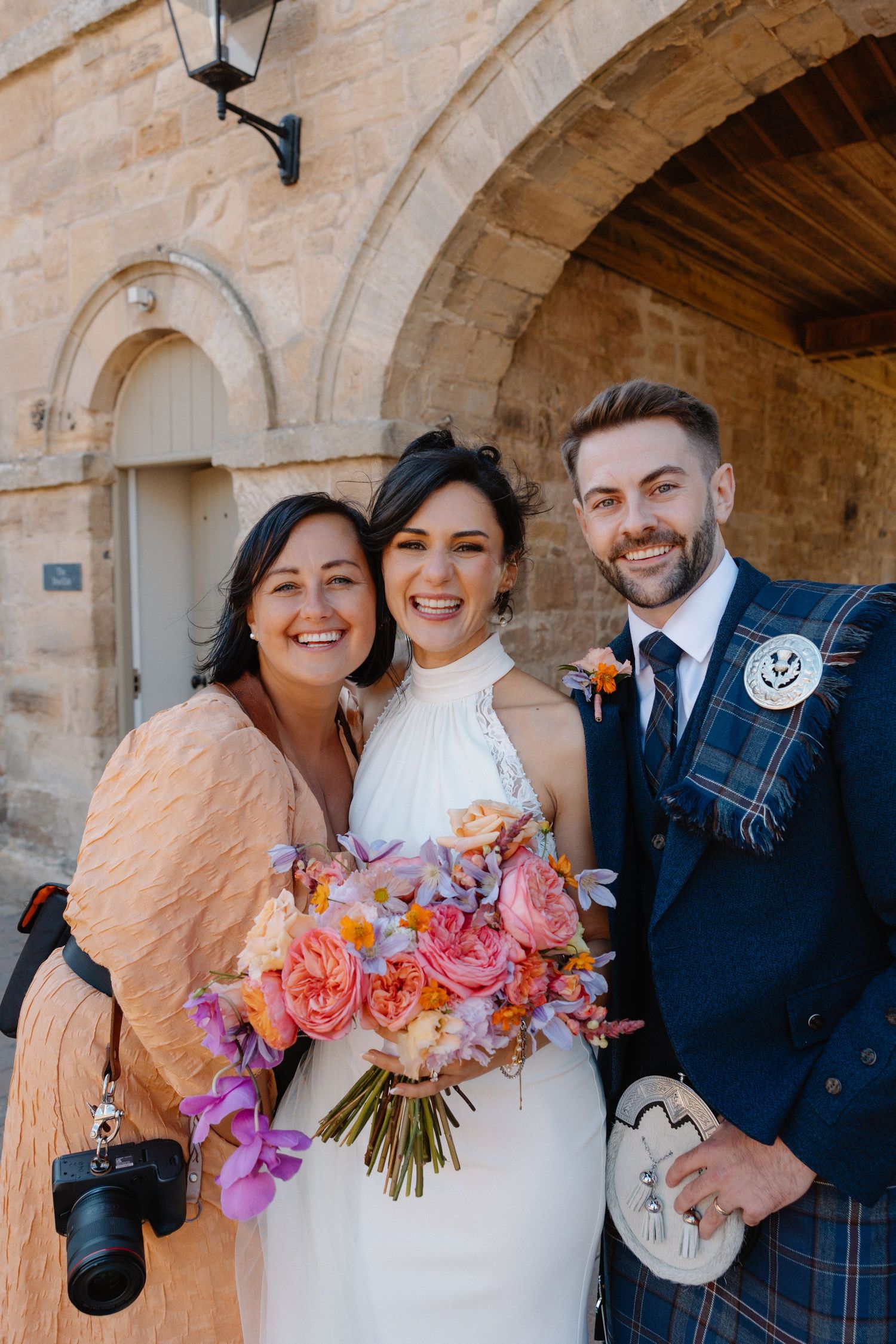 Bride in white halter dress holds colorful bouquet posing with groom in tartan kilt and female guest.