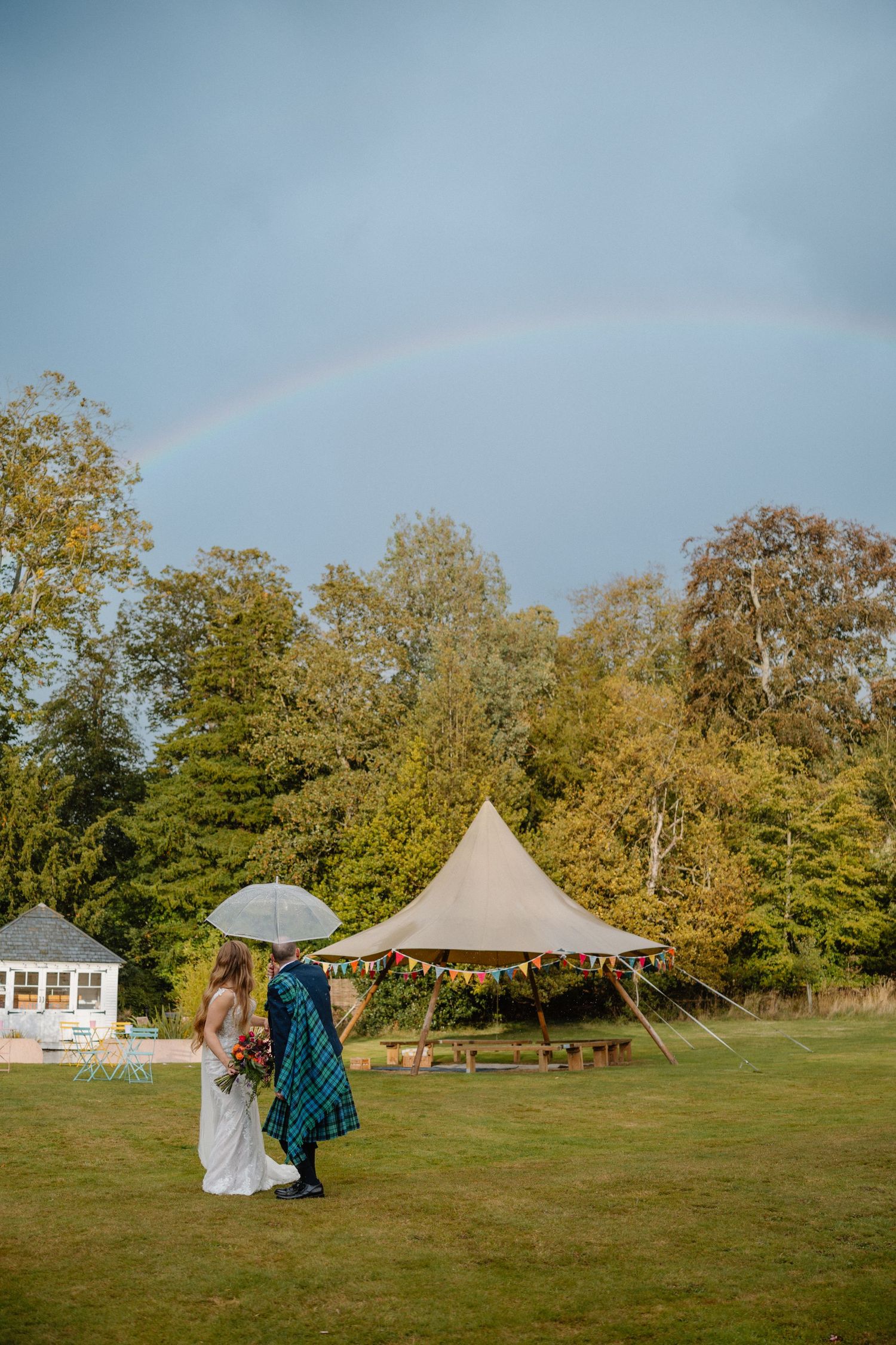 Bride and groom walk under umbrella with rainbow arching over a tipi wedding venue.