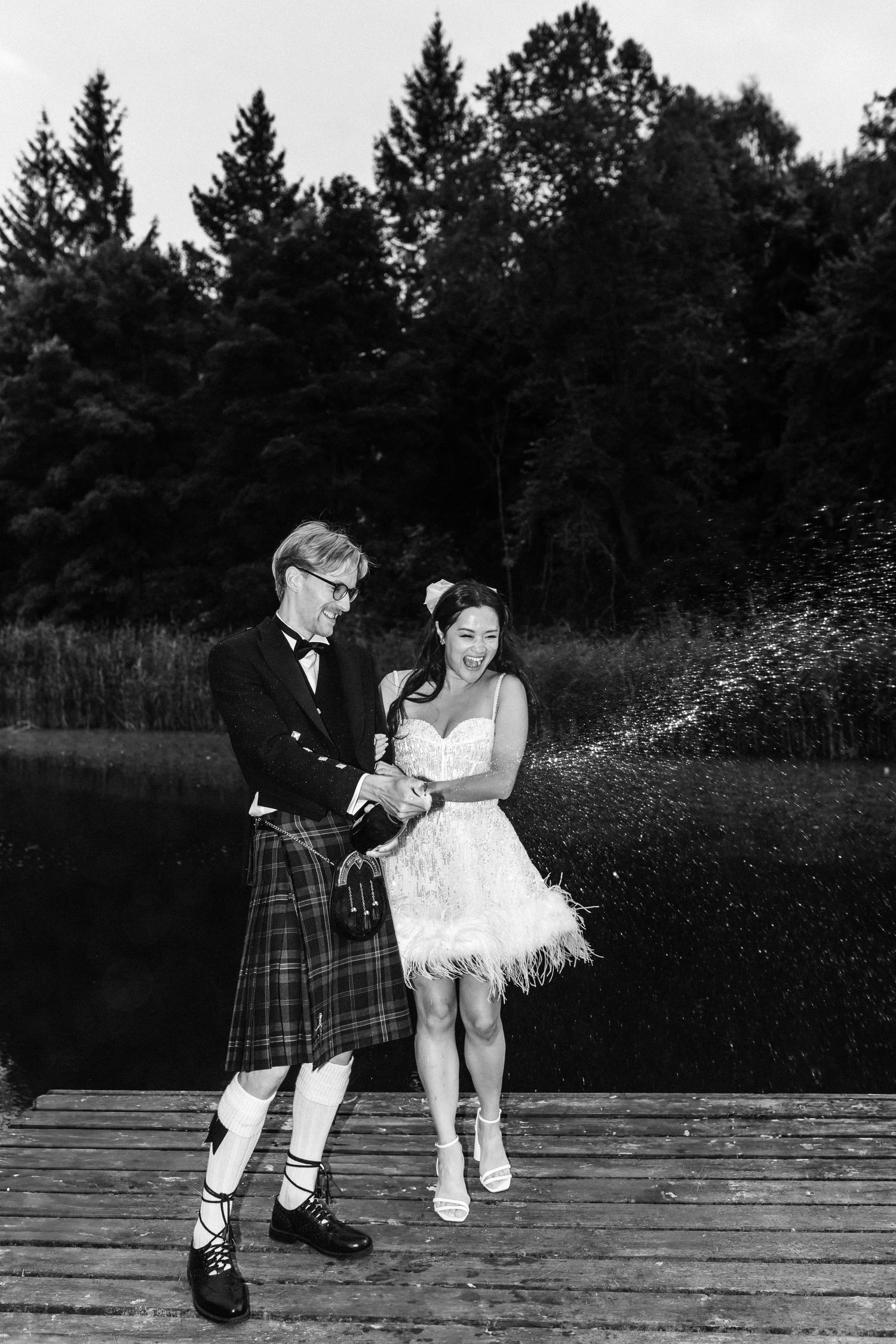Black and white photo of bride and groom popping champagne on a wooden dock by a lake.