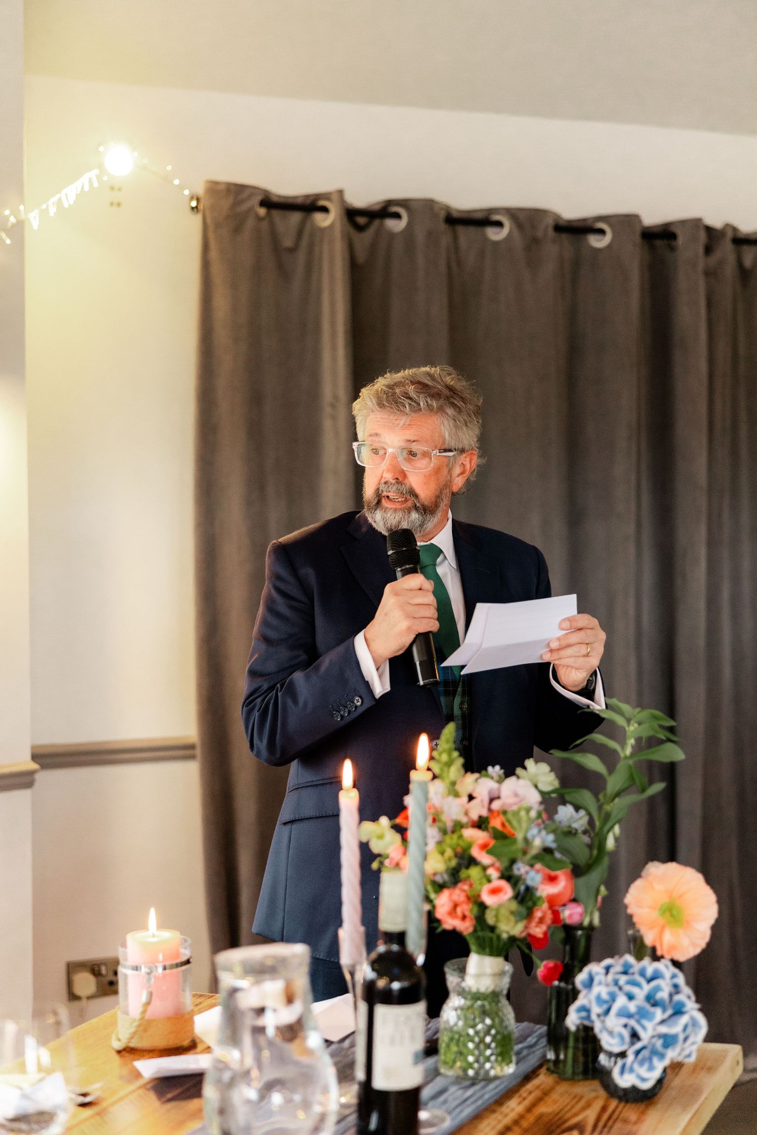 Bearded man in navy suit delivers speech holding microphone and paper at a wedding reception.