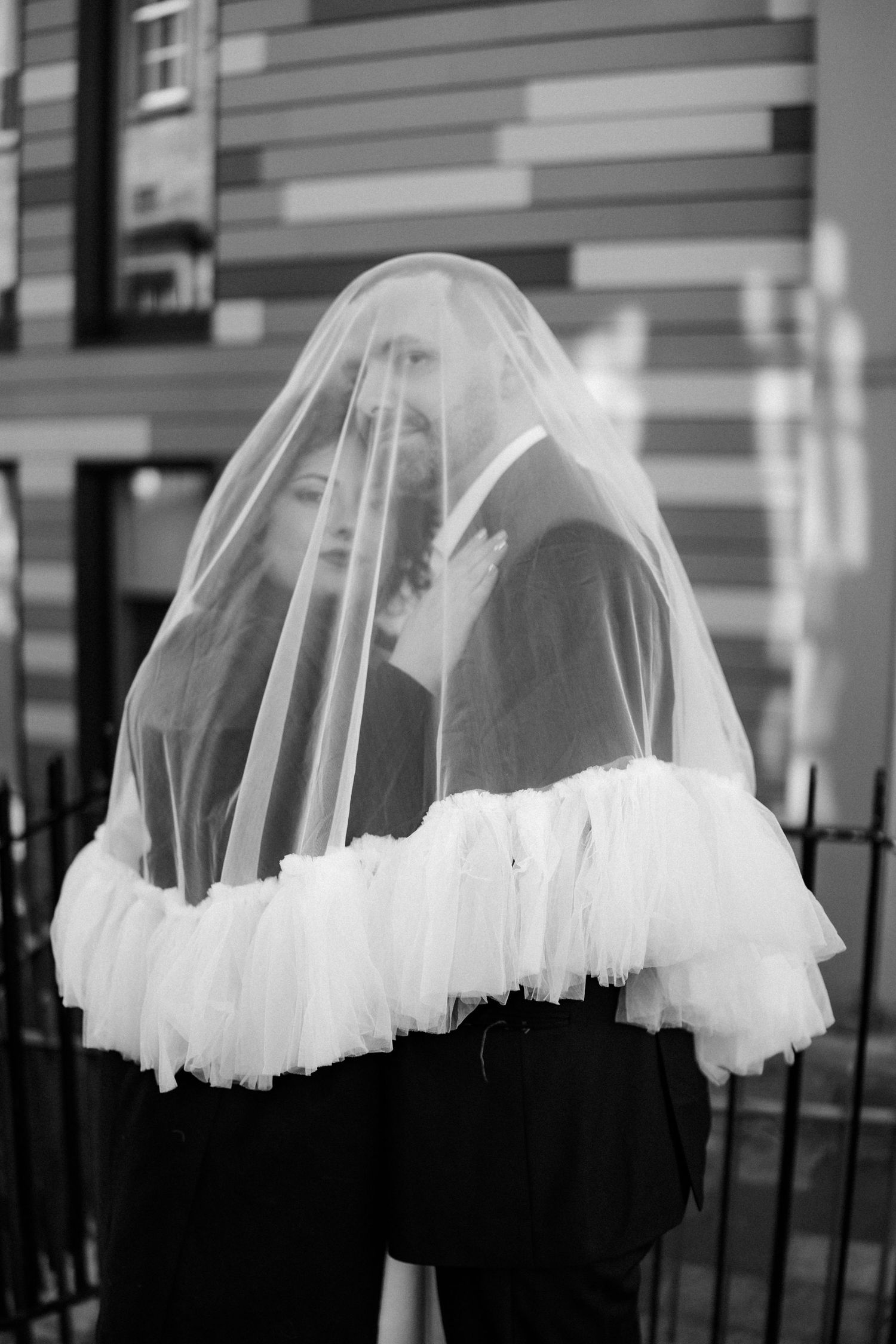 Black and white photo of a bride and groom sharing an intimate moment under a flowing wedding veil outdoors.