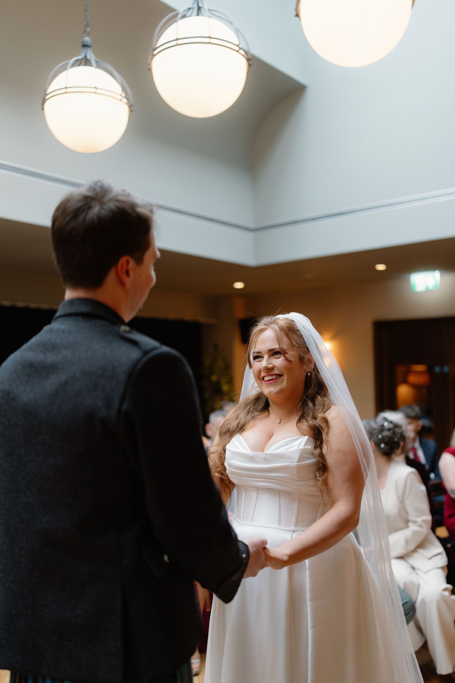 Bride in white satin gown holding hands with groom during indoor wedding ceremony.