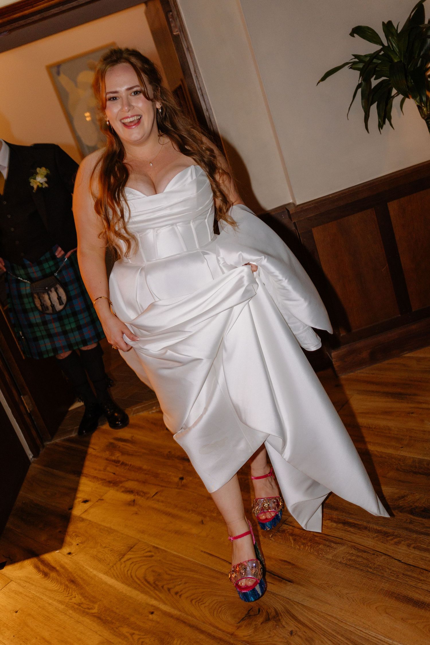 Smiling bride in strapless white satin wedding dress showing off colorful embellished heels on wooden floor.
