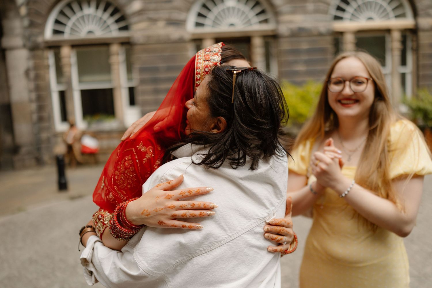 A bride in red traditional attire hugs a guest outdoors while a smiling woman in yellow dress looks on.