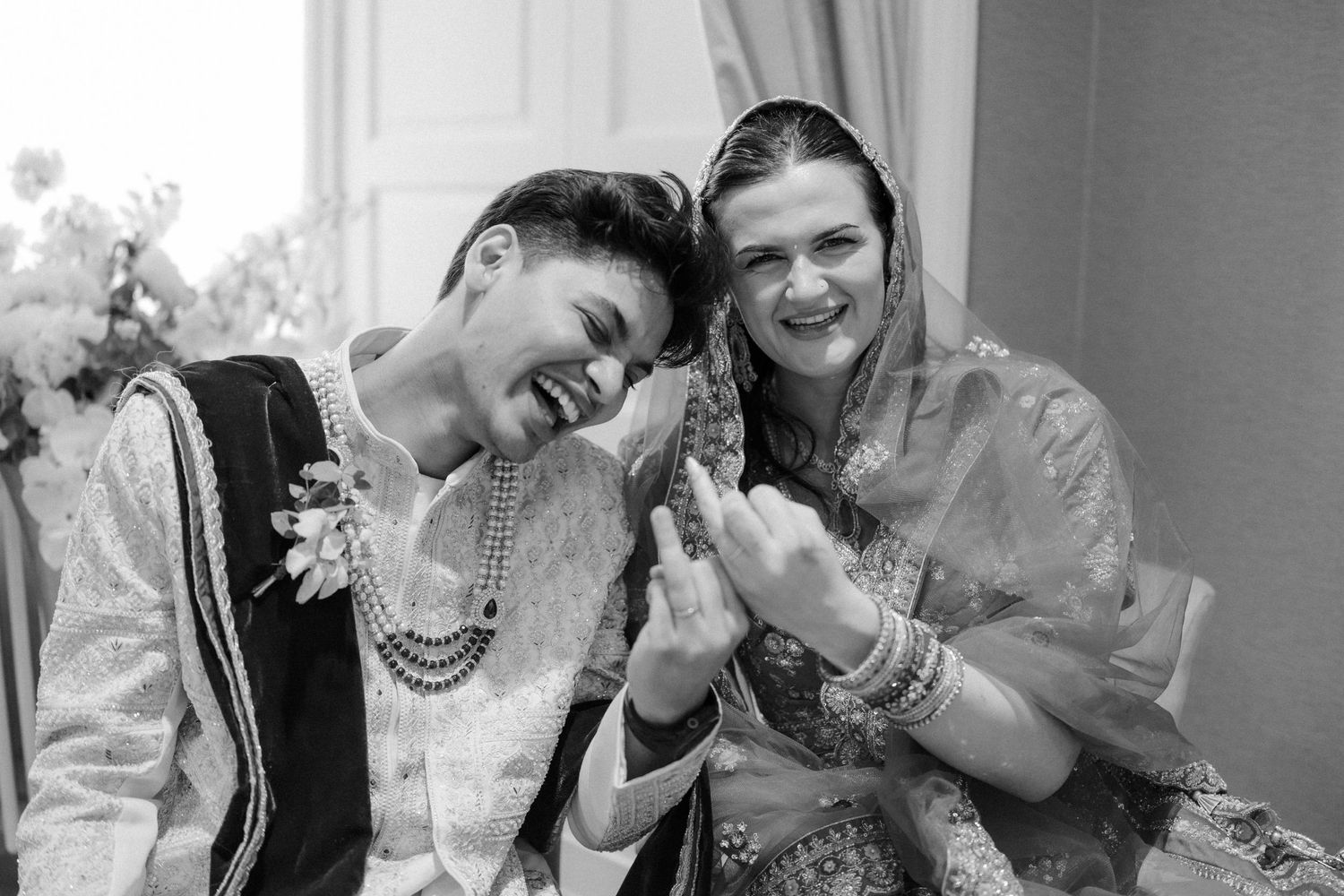 Two women in traditional South Asian attire laugh joyfully together in a black and white wedding photo.