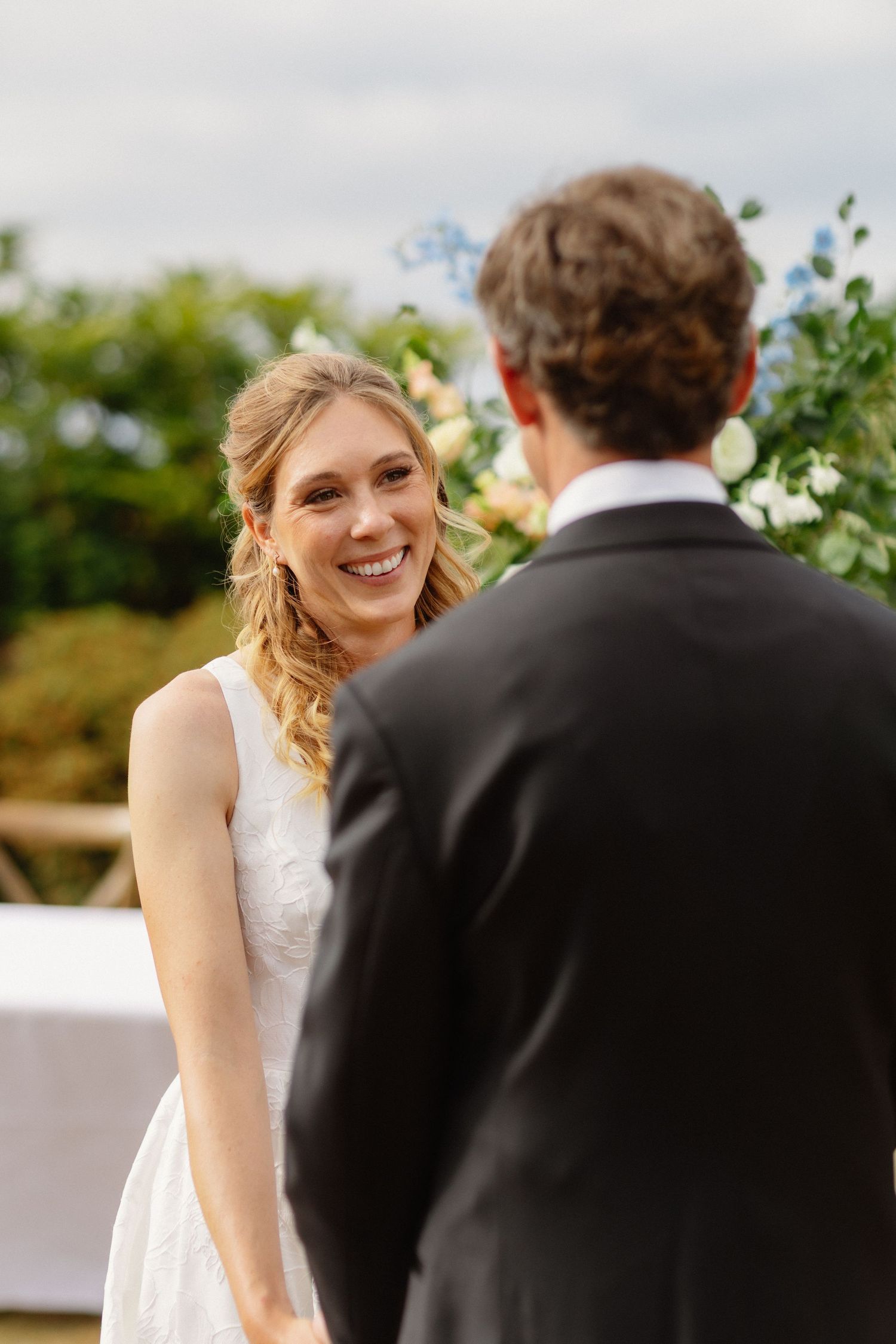 Smiling blonde bride in white dress holding hands with groom during outdoor wedding ceremony.