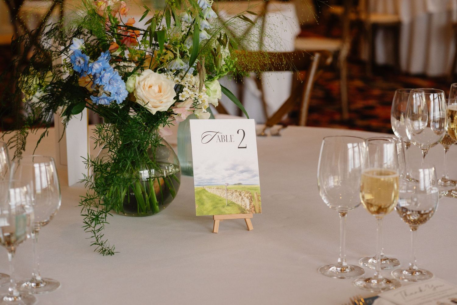 Elegant wedding reception table with floral centerpiece, wine glasses, and a painted Table 2 sign on a easel.