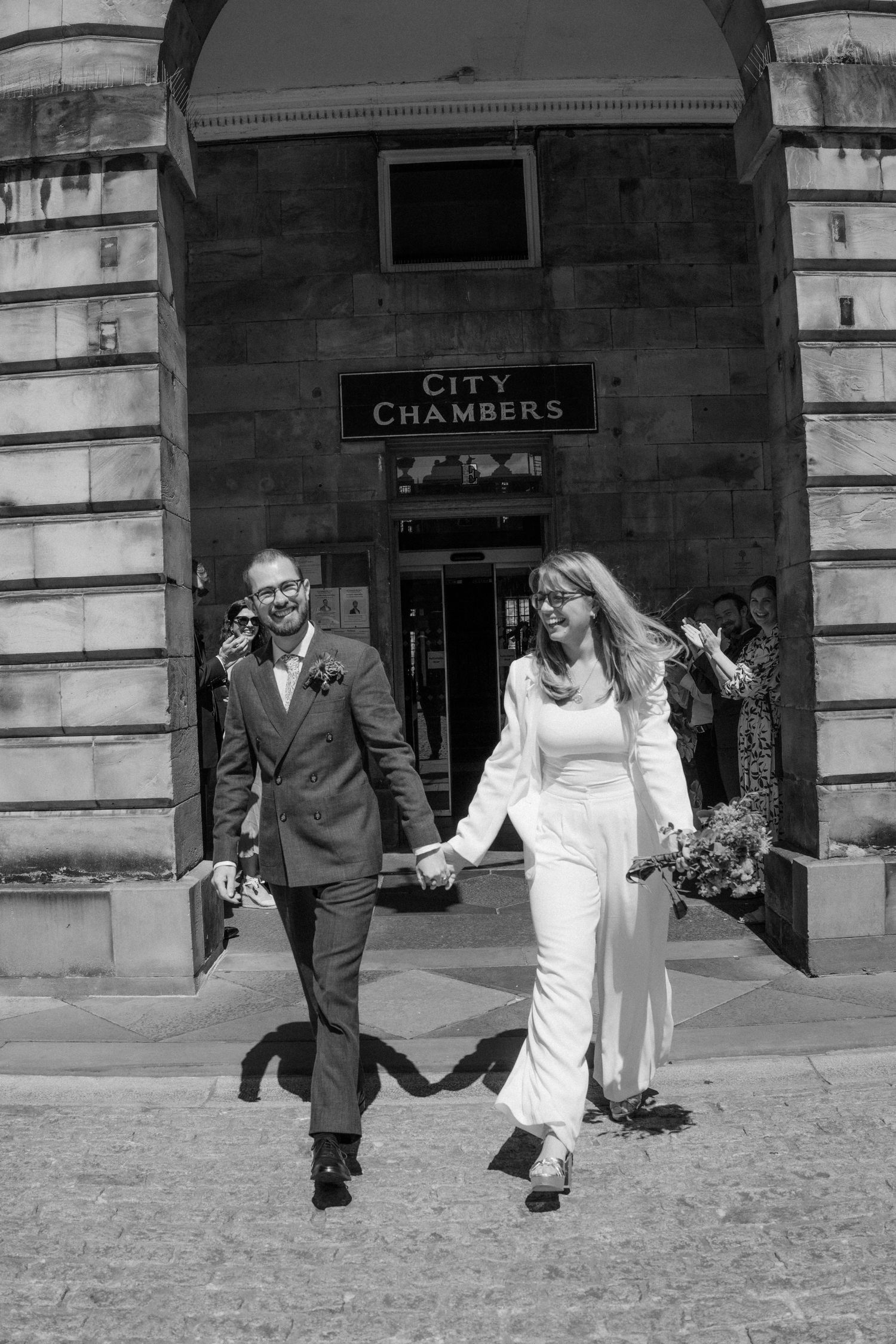 A couple walking and holding hands outside a historic city building in black and white photography.