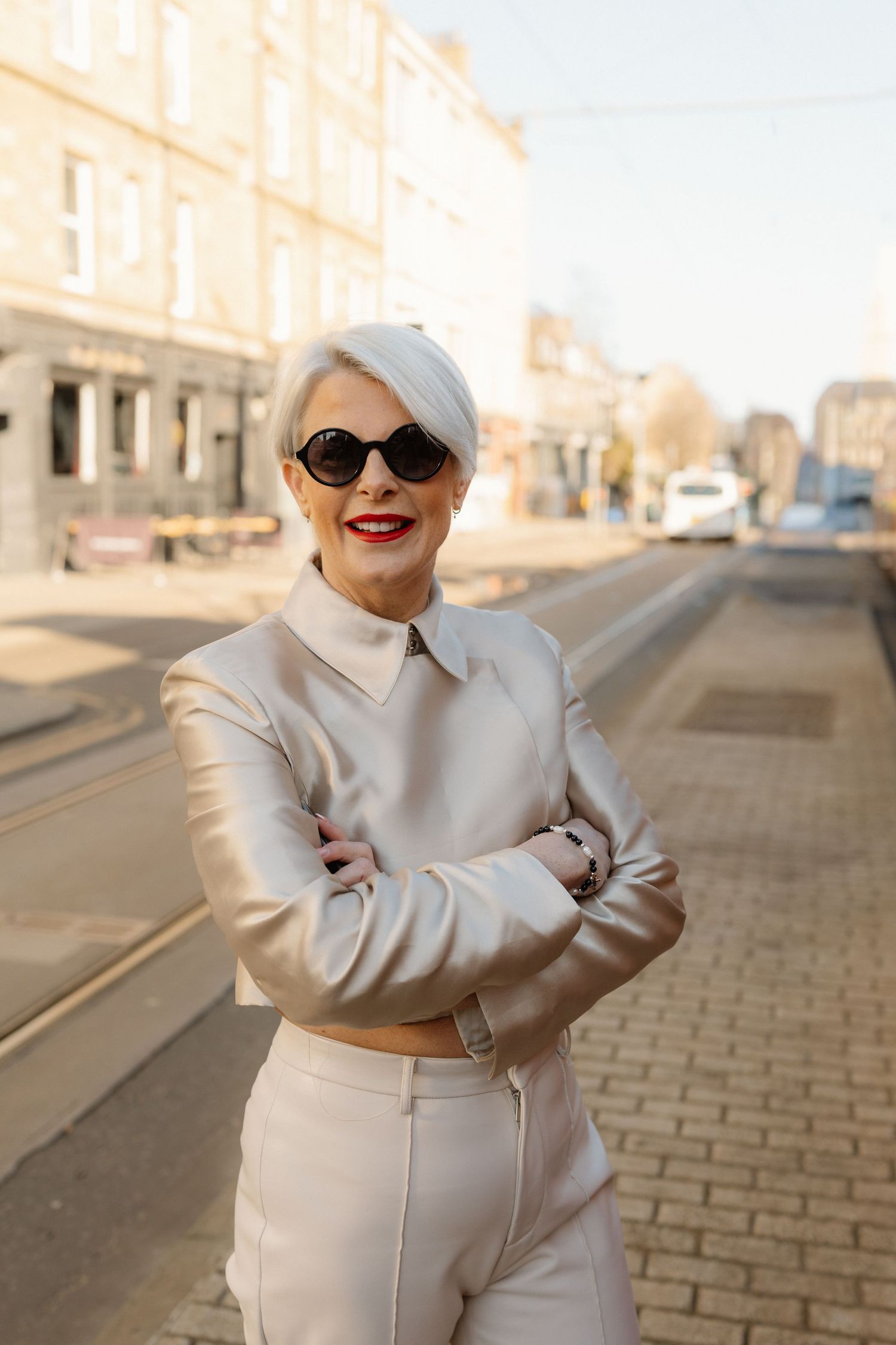 Stylish figure in cream loungewear and sunglasses poses confidently on cobblestone street at sunset.