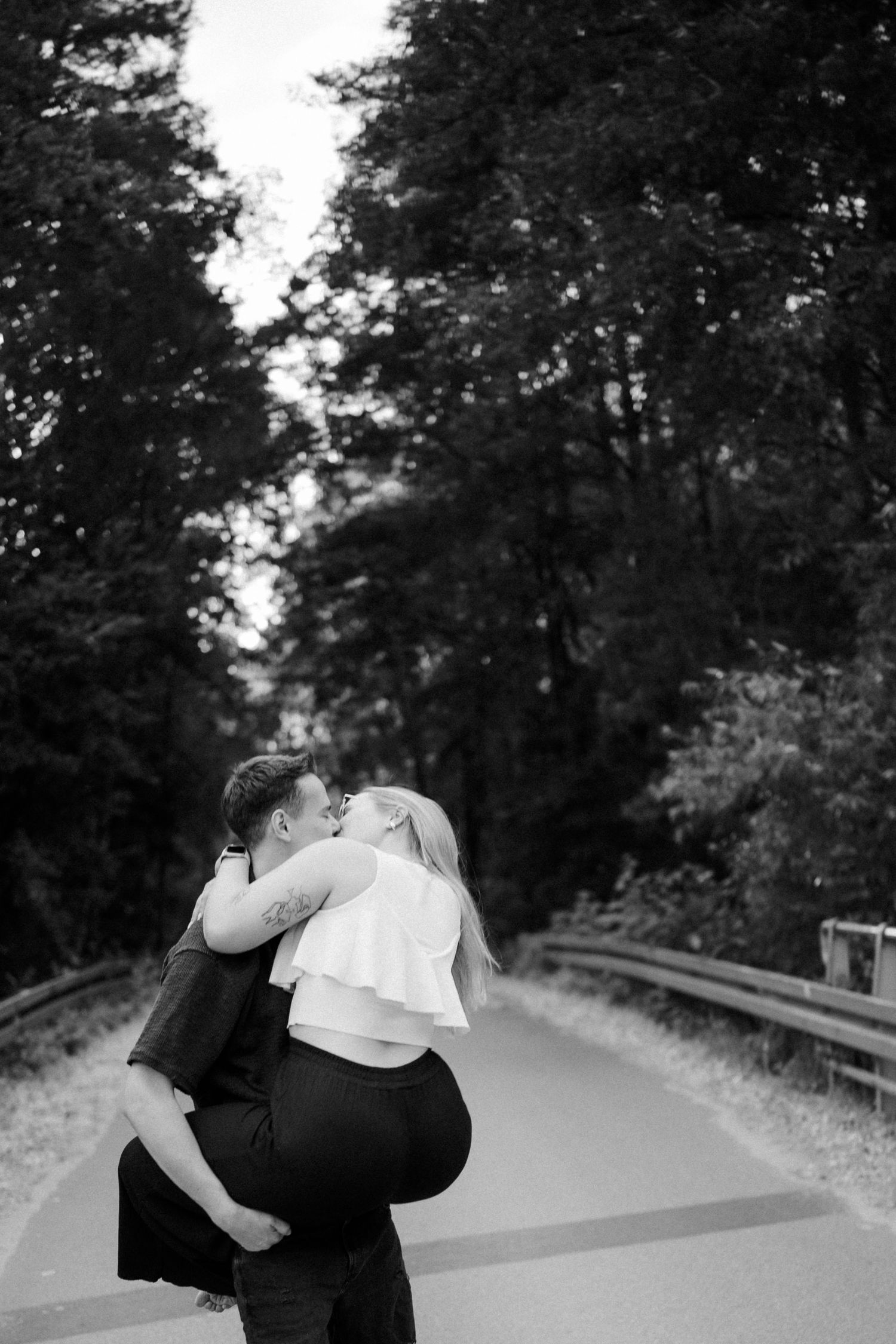 A couple embraces on a winding road surrounded by trees in dramatic black and white.