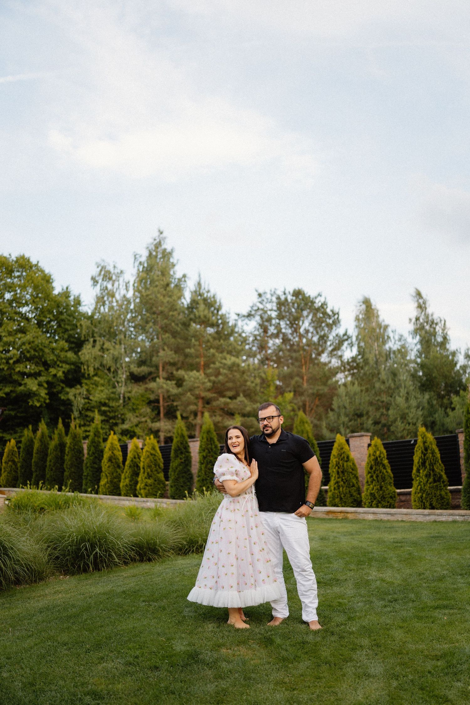 Outdoor garden photoshoot with couple on manicured lawn surrounded by evergreen trees.