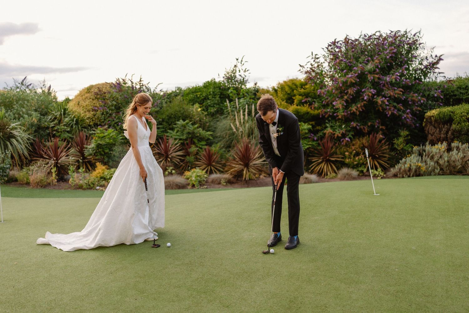 Bride and groom playing golf on a putting green in wedding attire, surrounded by lush garden greenery.