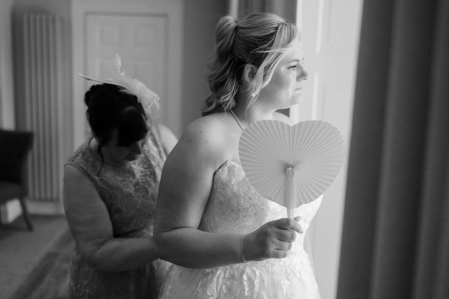 Black and white photo of a bride in lace gown holding a fan near window, with bridesmaid adjusting her dress.