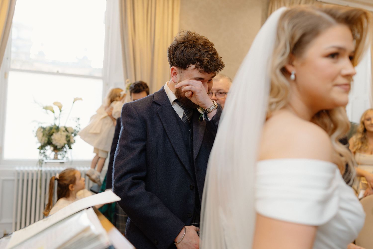 Emotional groom wiping tears as bride stands beside him during heartfelt wedding ceremony.